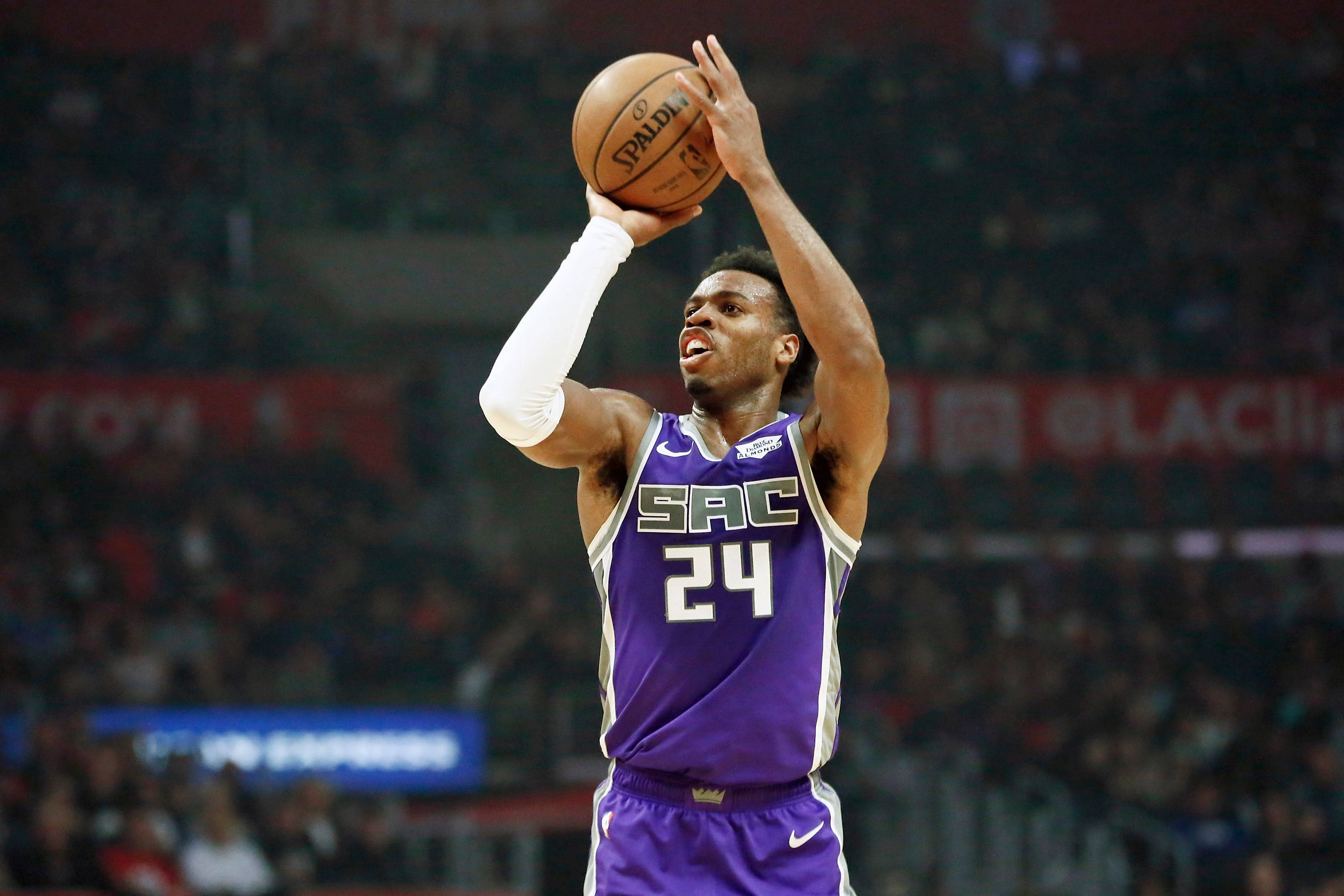 薩克拉門托國王隊 Buddy Hield 擊球 壁紙 — 一名身穿紫色薩克拉門托帝王隊球衣的籃球運動員在場館燈光下專注地投擲Spalding籃球。