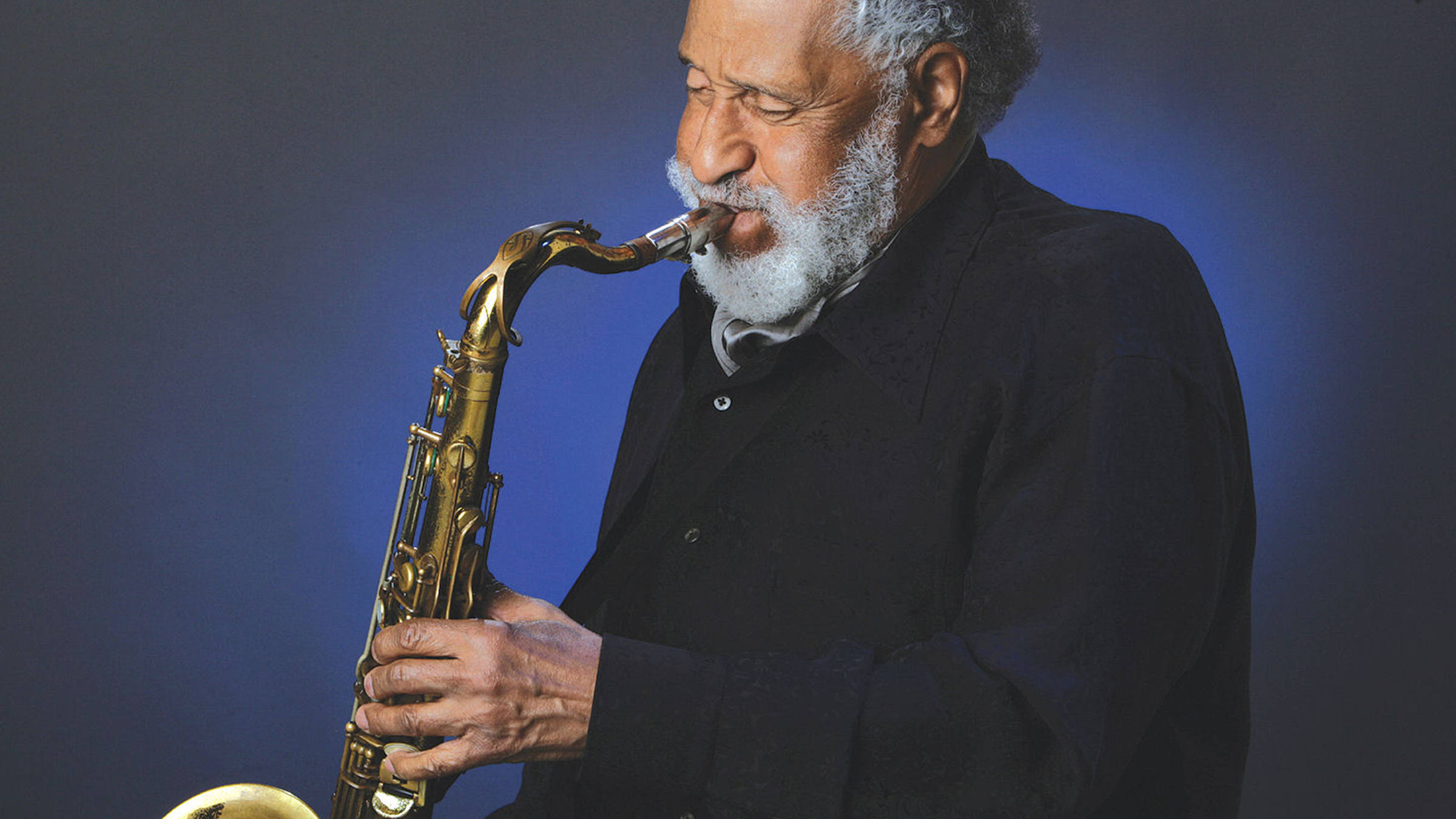 薩克斯風巨匠 Sonny Rollins 壁紙 — 一位留著白鬍子的長者，在深藍色工作室背景下吹奏金色色士風。
