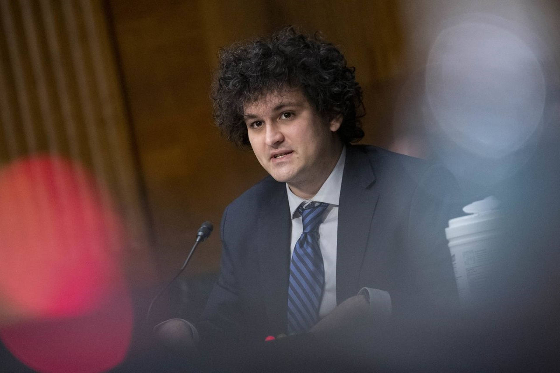 身穿西裝打領帶的男人在正式聽證會上發言，前景有模糊的燈光。