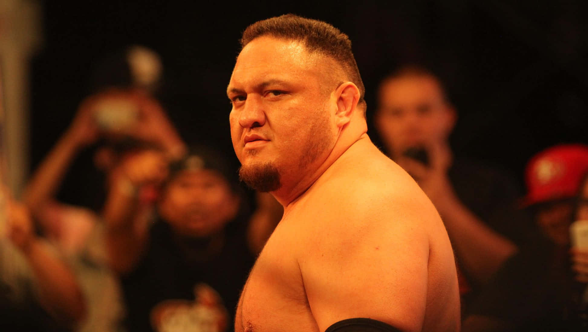 薩摩亞·喬WWE Raw賽事中的側臉反應 壁紙 — 一位肌肉發達的摔角手表情嚴肅地站在燈光昏暗的競技場中，粉絲們用手機捕捉這一刻。