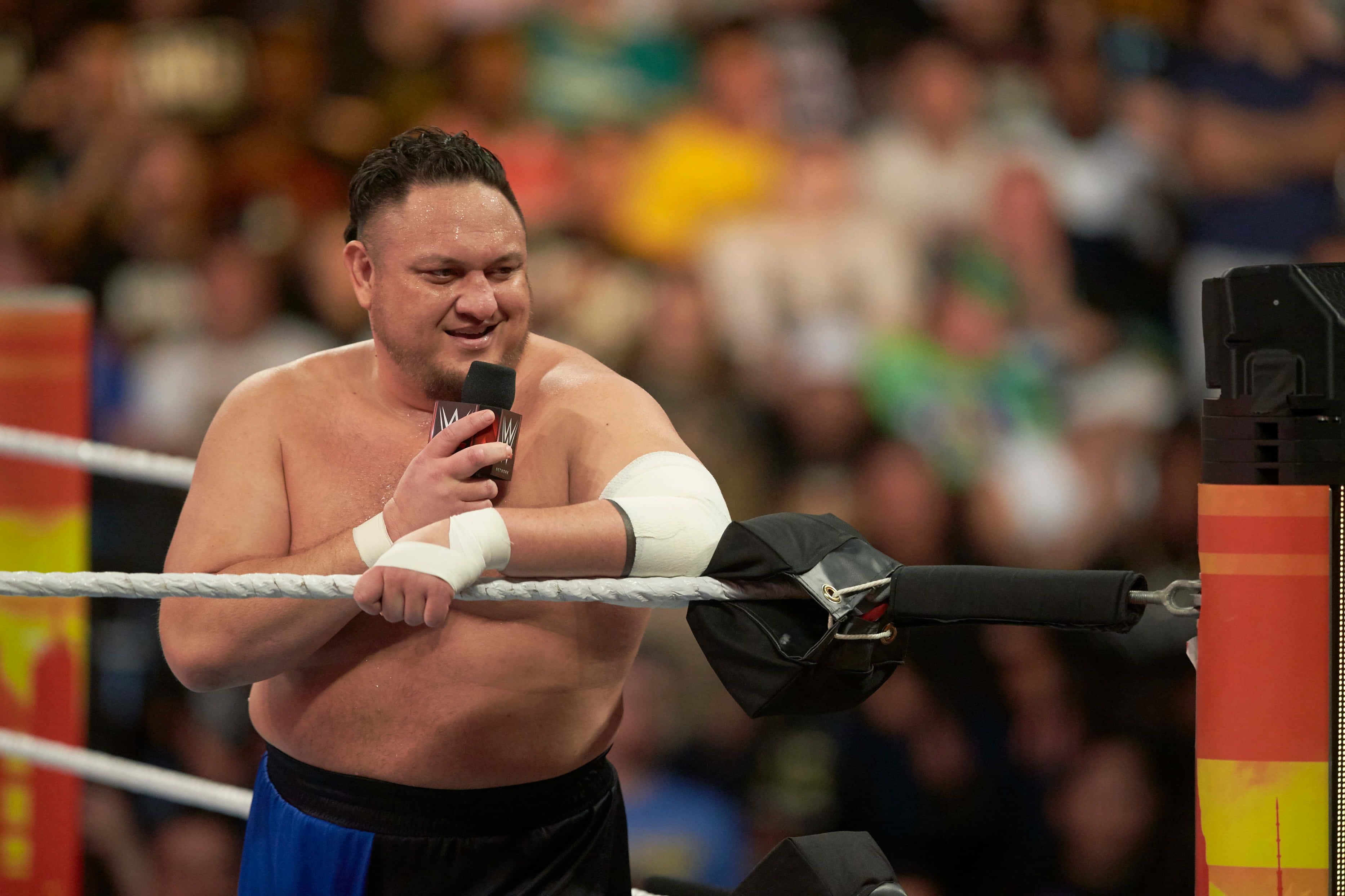薩摩亞·喬在NXT Takeover 36賽事中談話 壁紙 — 一位肌肉發達的摔角手穿著藍色短褲，手持麥克風，靠在擂台繩索上，對著人群微笑。