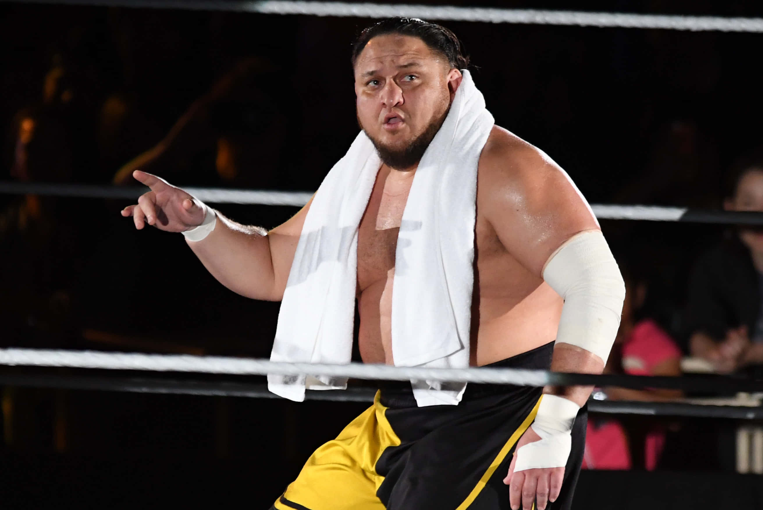 薩摩亞·喬於WWE Live東京賽事入場 壁紙 — 一位肌肉發達的摔角手穿著黑色和黃色短褲，肩上披著白色毛巾，在明亮的擂台上激烈地指著。