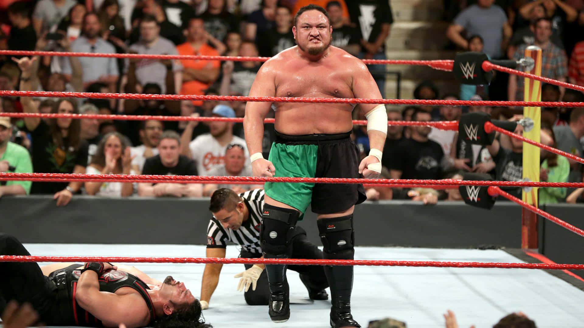 薩摩亞·喬於WWE Payback 2017擊敗賽斯·羅林斯 壁紙 — 一位肌肉發達的摔角手穿著綠色和黑色的短褲，在WWE擂台上勝利地站著，而他的對手則被打敗躺在地上，裁判正在檢查他。