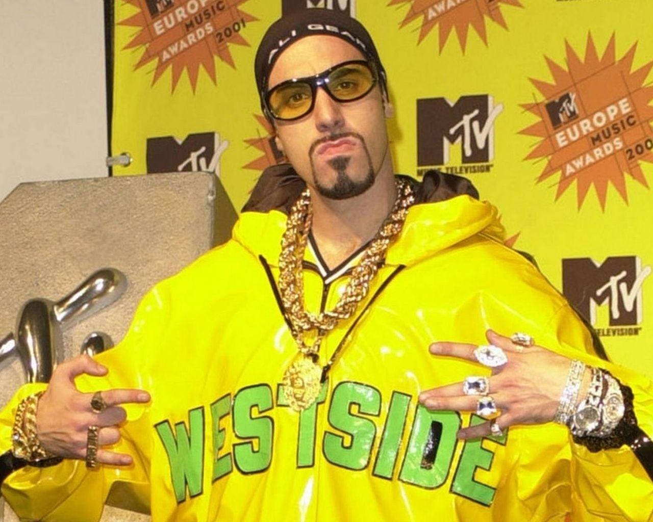 薩沙·巴隆·科恩 飾演電視節目《Ali G》中的角色 壁紙 — 一名身穿亮黃色 'WESTSIDE' 連帽衫、佩戴金鏈的男子在2001年MTV歐洲音樂大獎上自信地擺姿勢。
