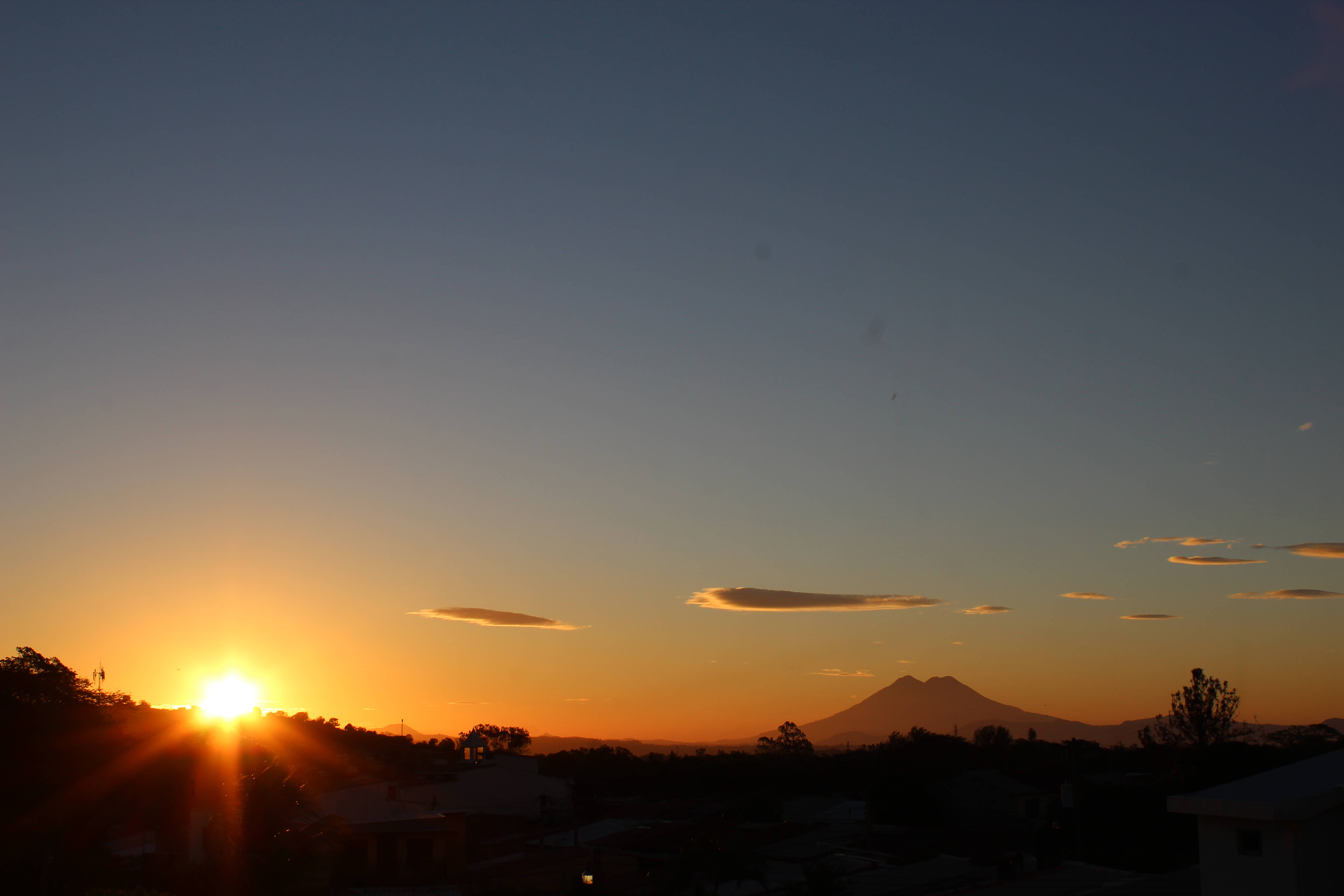 薩爾瓦多日落風景 壁紙 — 夕陽在遠山後方閃耀，剪影般的屋頂和樹木勾勒出地平線。