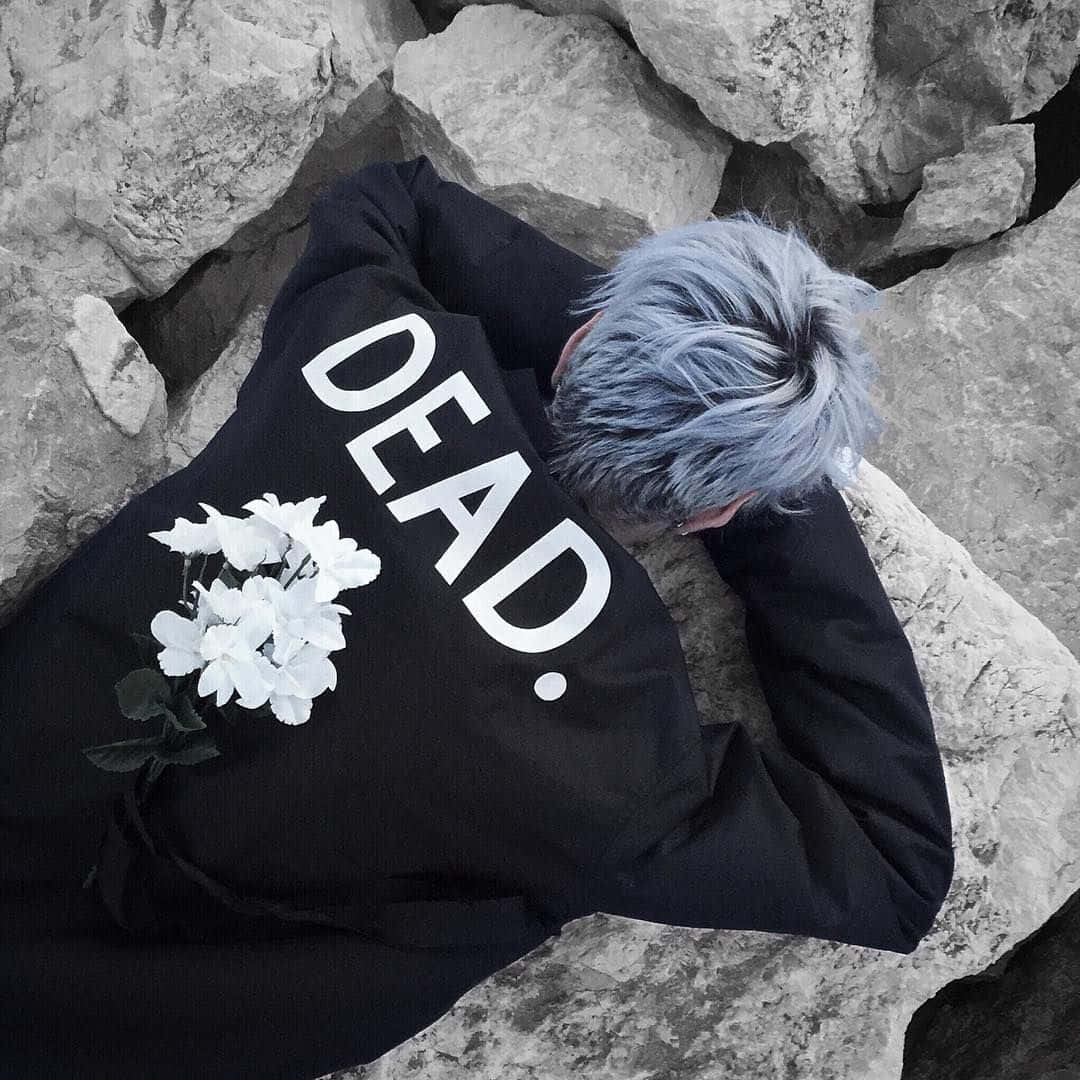 藍色頭髮的人躺在岩石上，穿著黑色連帽衫，上面有「DEAD.」和白色花朵。