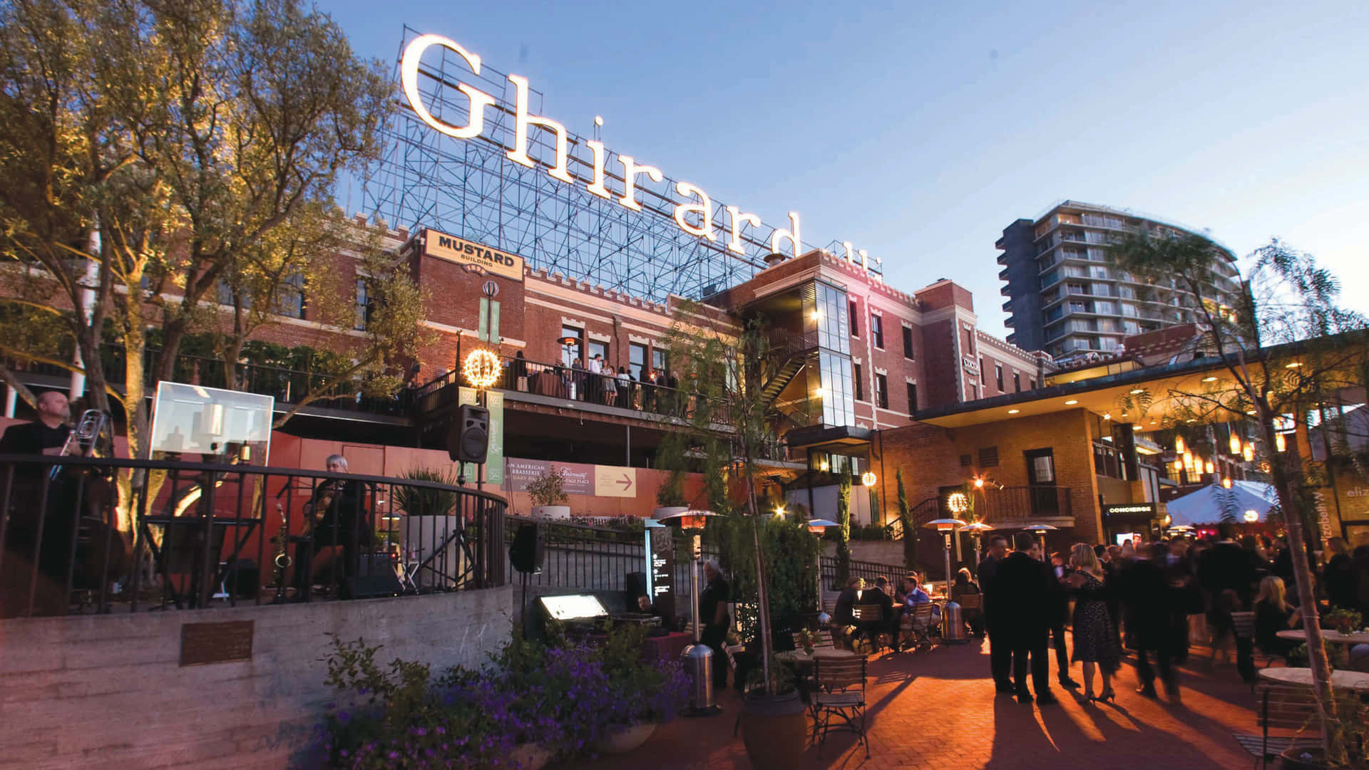 藍天下的吉拉德利廣場 壁紙 — Ghirardelli Square 一個熱鬧的戶外庭院，沐浴在溫暖的晚光中，人們在發光的串燈下用餐和散步。