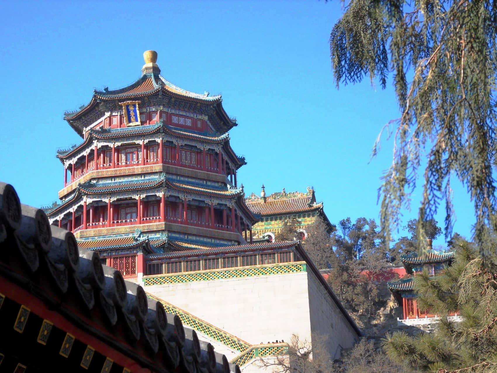 藍天下的夏日宮殿 壁紙 — 一座宏偉的多層中式寶塔，紅色柱子和藍色瓦片屋頂，在樹枝環繞的清澈藍天下聳立。