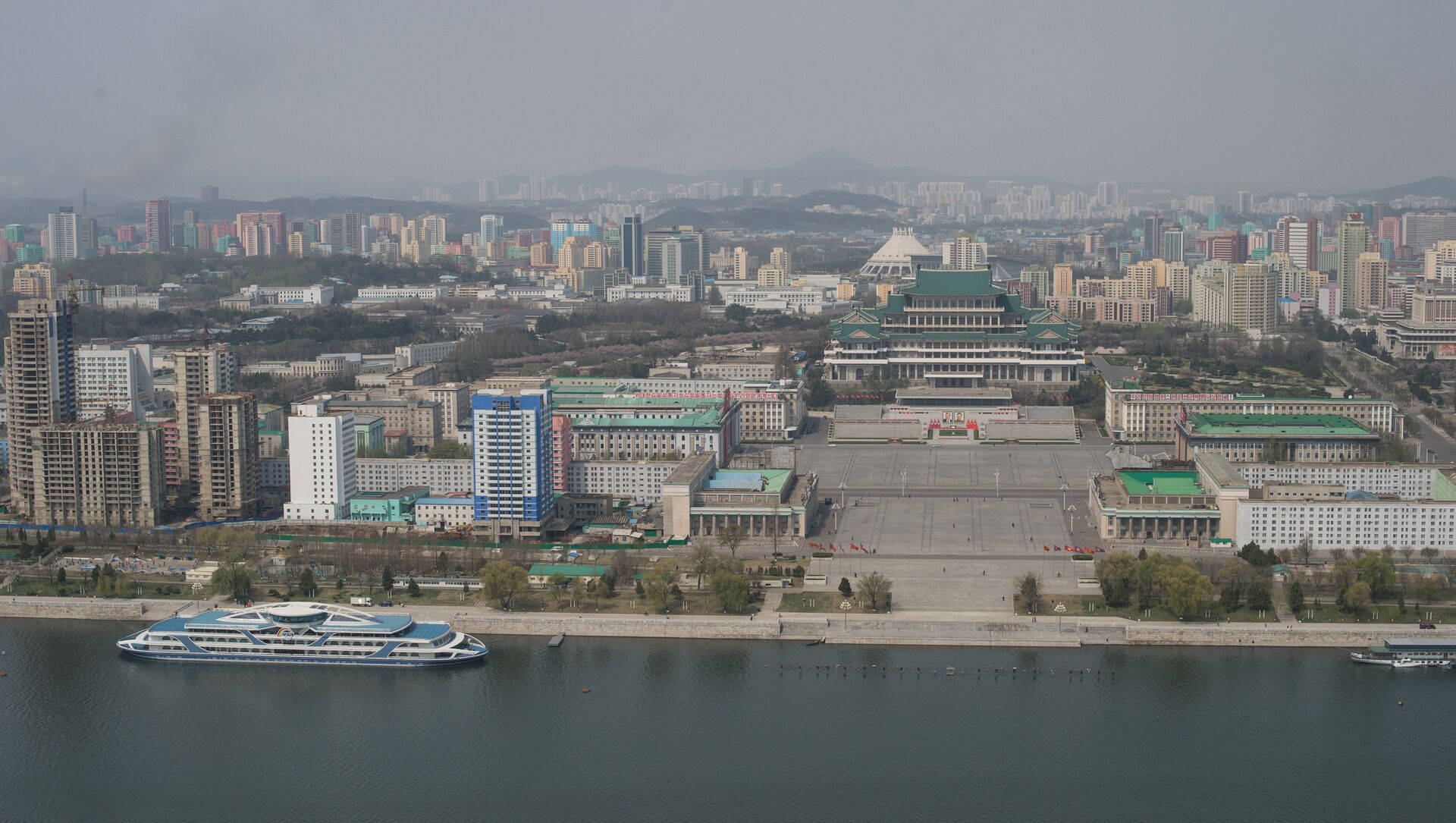 一條寬闊的河流上有一艘船，經過一個建築密集的城市，天空朦朧。