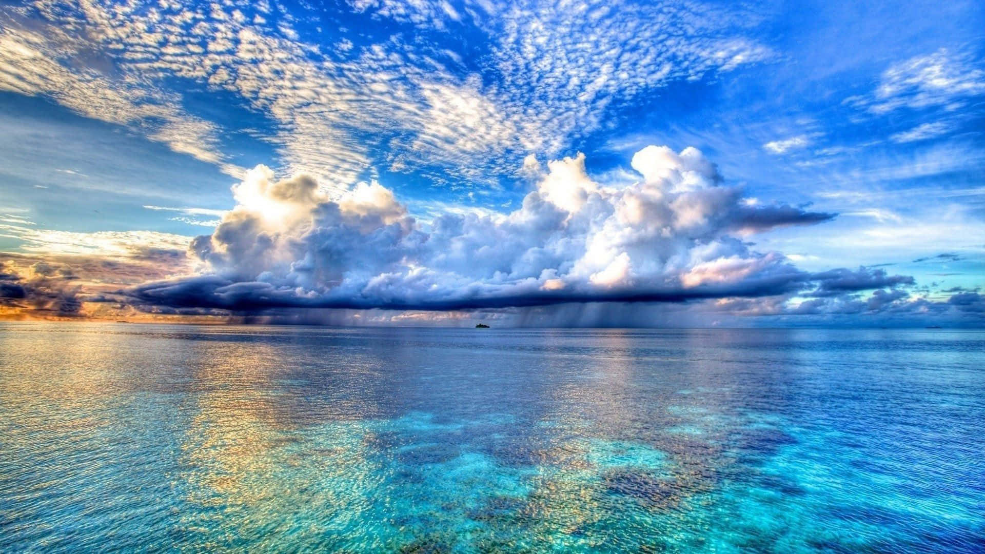 藍天海灘主題背景 壁紙 — 廣闊的藍綠色海洋與戲劇性的天空相遇，陽光照射的雲層和遠處的陣雨籠罩在平靜的水面上。
