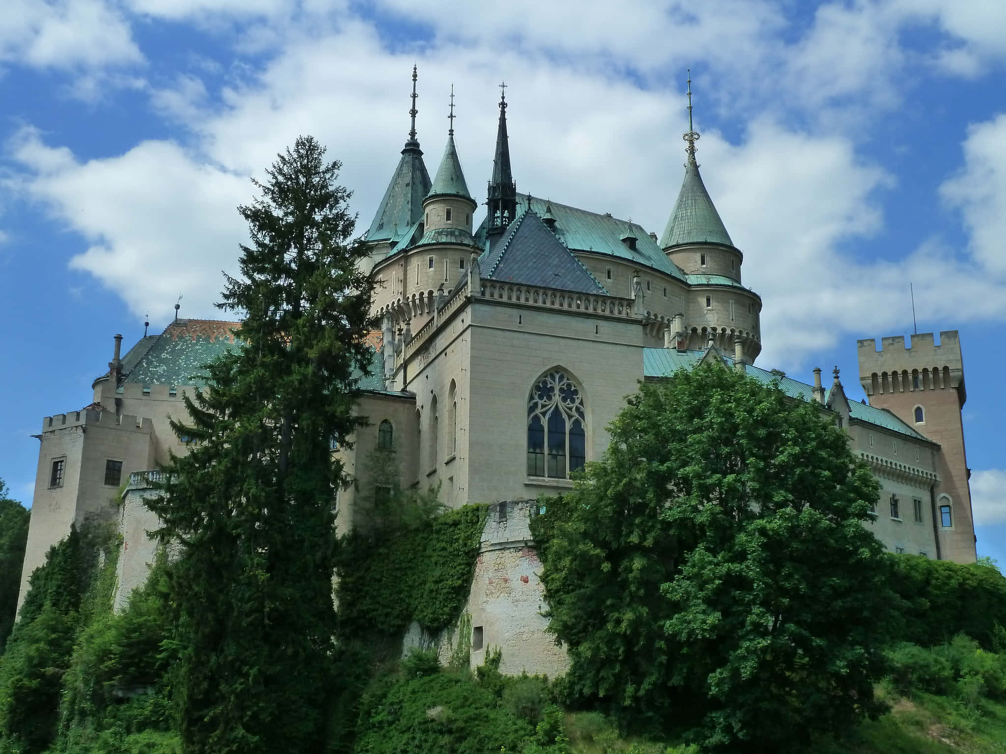 藍天背景下的波亞尼奇城堡 壁紙 — 一座宏偉的哥德式城堡，擁有綠色銅屋頂和石塔，在明亮的藍天和蓬鬆的白雲下，雄偉地聳立在茂密的綠樹之上。