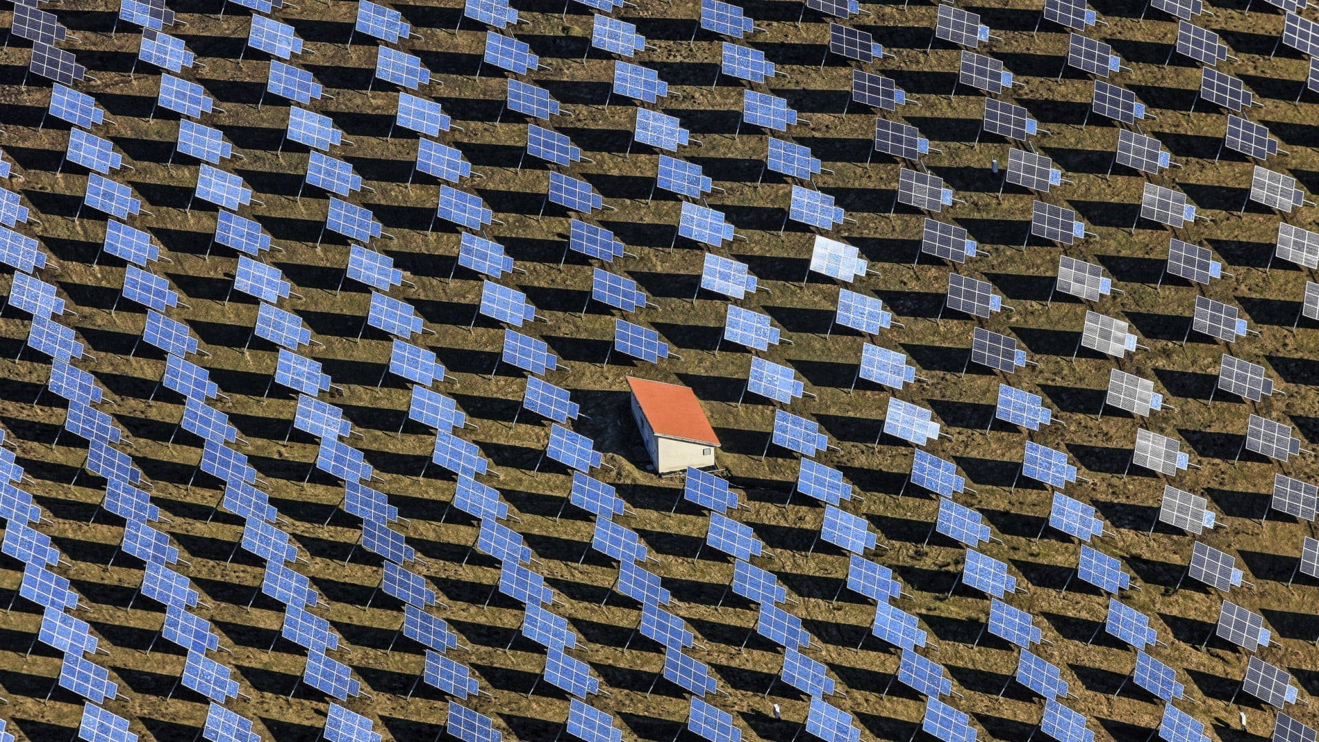 一座紅色屋頂的小建築，坐落在田野中的太陽能板行列之間。