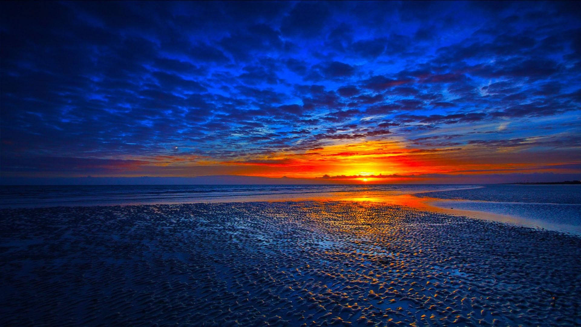藍橙夕照美景 壁紙 — 戲劇性的日落將天空染成火熱的橙色和深藍色，金色的光線反射在紋理濕潤的海灘上。
