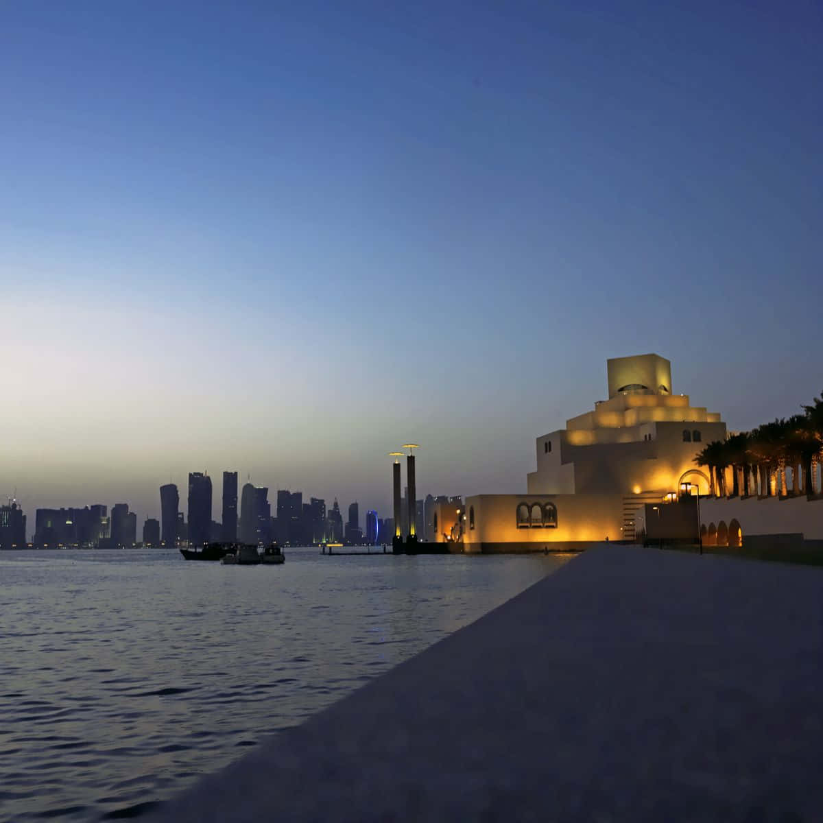 藍橙色調的伊斯蘭藝術博物館廣場夜景 壁紙 — 一座現代博物館在黃昏時分散發著溫暖的光芒，旁邊是平靜的水面，背景是城市天際線。