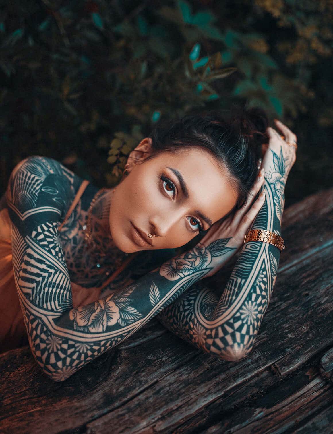 藍紫髮色的紋身女孩凝望遠方 壁紙 — 一位紋身黑髮女子將頭靠在手臂上，置於風化木頭上，周圍環繞著茂密的綠色樹葉。
