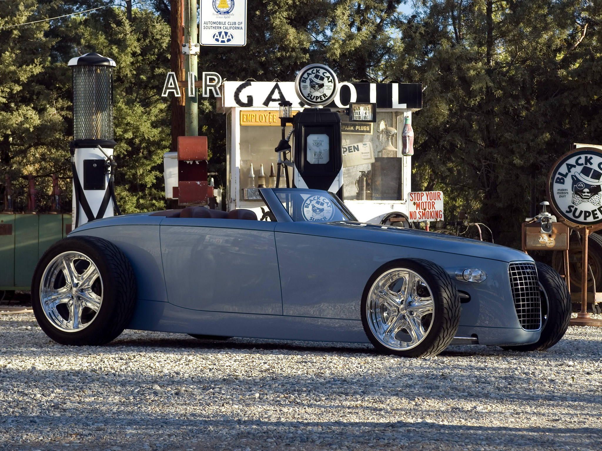 藍色 Caresto Volvo iPhone 壁紙 — 一架時尚的銀色定制熱棒車配備鍍鉻輪圈，停泊在碎石地上，前方是帶有復古標誌的懷舊加油站。