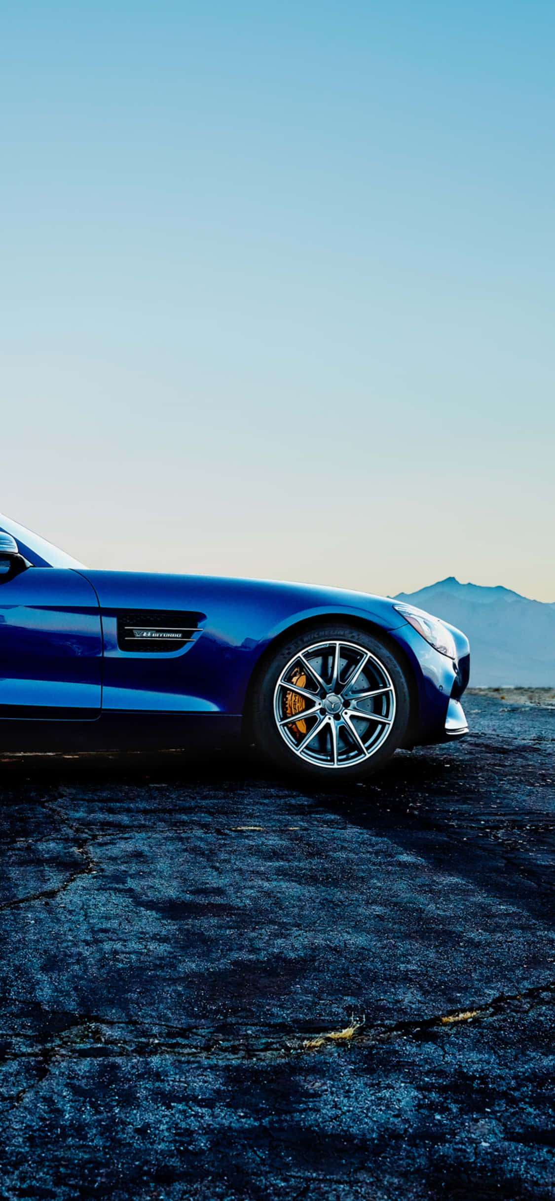 藍色 iPhone XS Mercedes AMG 背景 壁紙 — 一架時尚的藍色跑車停泊在裂開的瀝青路上，遠山在晴朗的天空下。