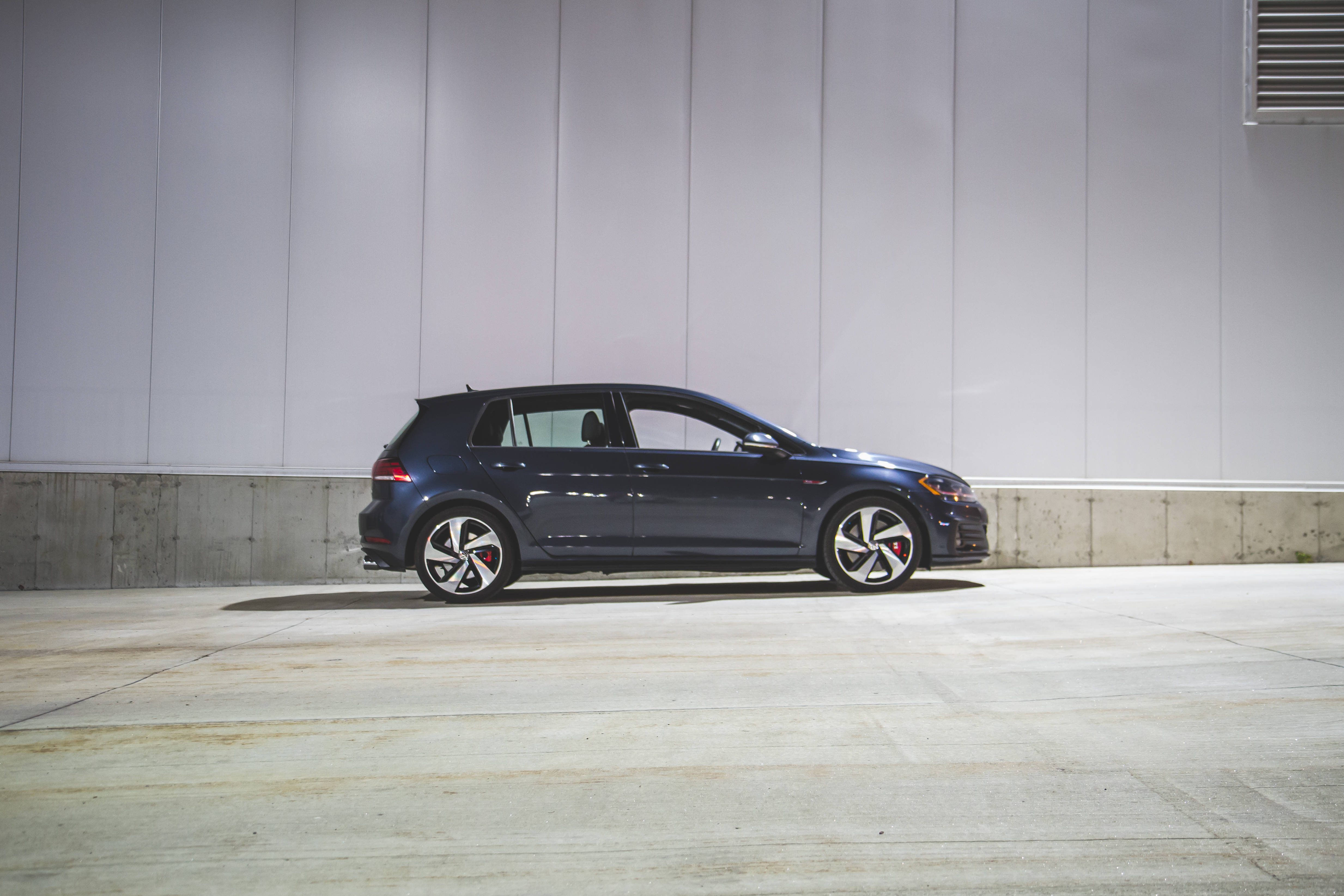 藍色 Volkswagen Golf GTI 側面視角 壁紙 — 一架時尚深藍色運動掀背車停泊在簡約混凝土車庫，背景為純白牆壁。