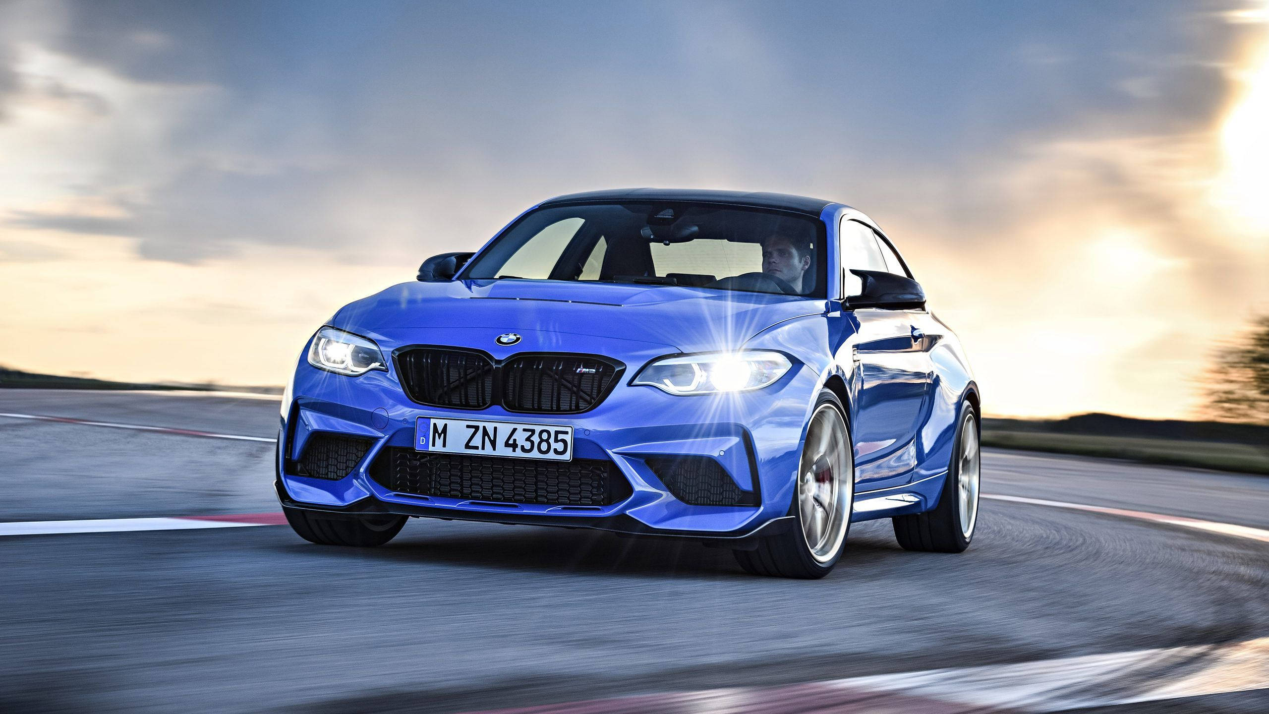 藍色BMW高速駛過彎道 壁紙 — 一輛充滿活力的藍色BMW M2 Coupe在日落時分沿著賽道疾馳，車頭燈閃爍，帶有動態模糊效果。