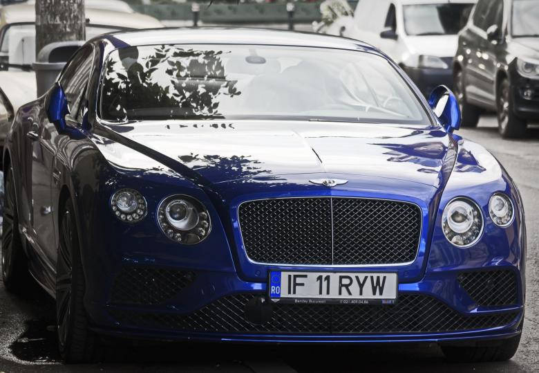 一輛藍色 Bentley Continental GT 停泊在濕漉漉的城市街道上，帶有樹木倒影。