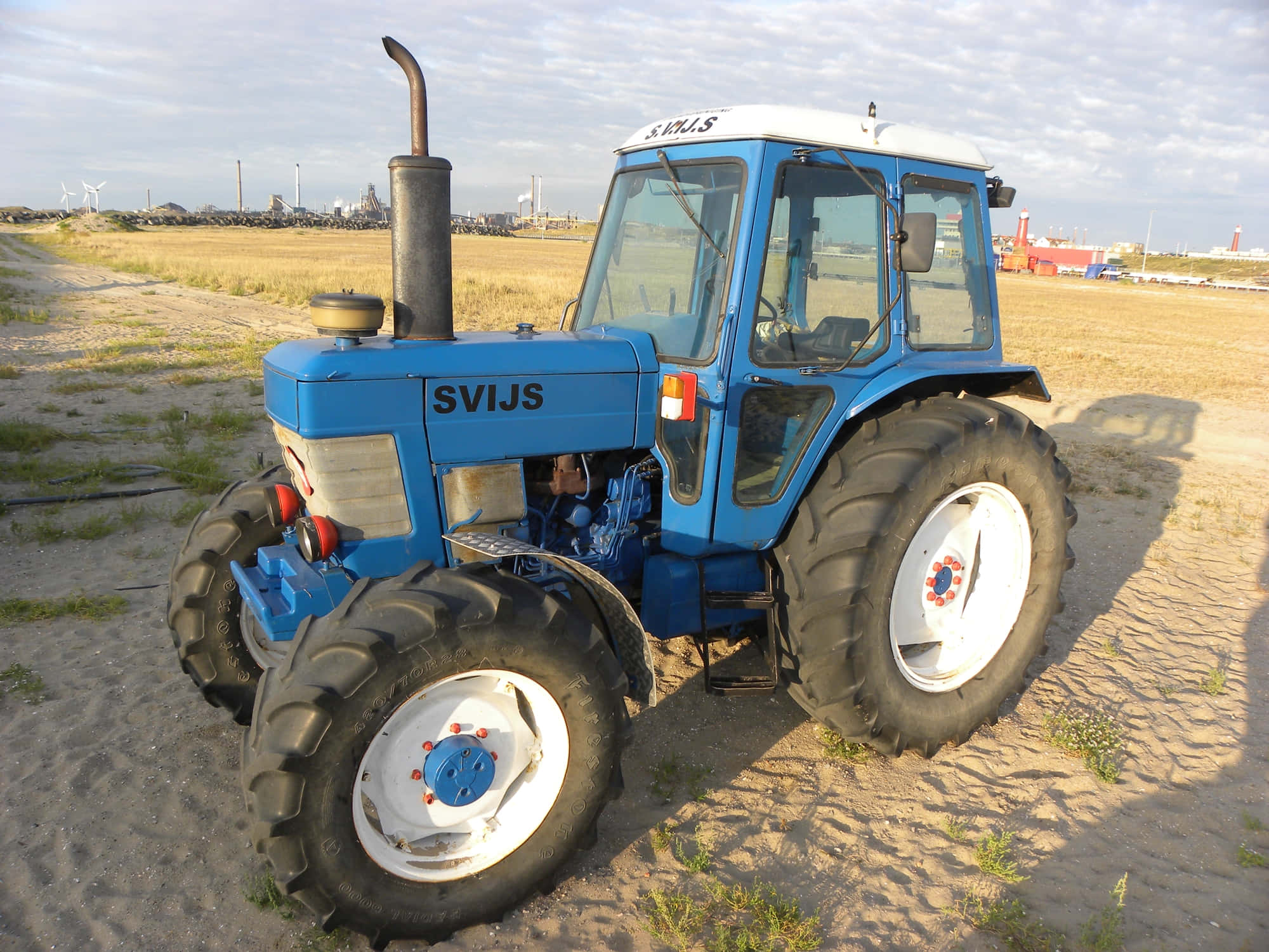 藍色Ford 6710拖拉機 壁紙 — 一輛白色車輪的亮藍色SVIJS拖拉機停泊在沙地上，天空多雲，遠處有工業建築。