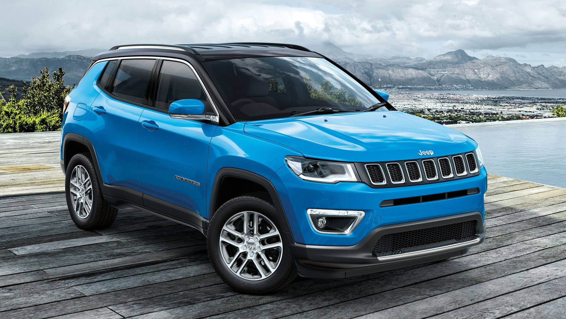 藍色Jeep Compass SUV停在木甲板上，背景是沿海城市和山脈。