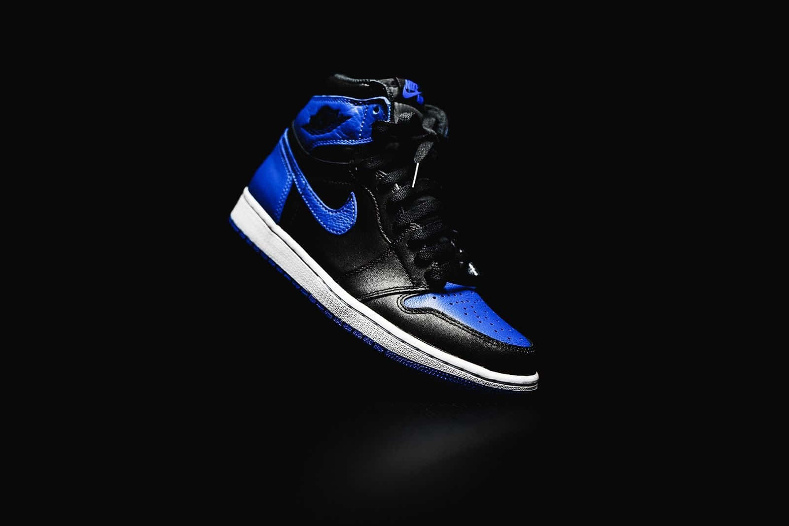 藍色Jordan Nike AJ One Retro Royal 壁紙 — 一雙時尚的黑色和皇家藍色高筒運動鞋在純黑色背景上漂浮，帶有戲劇性的燈光。