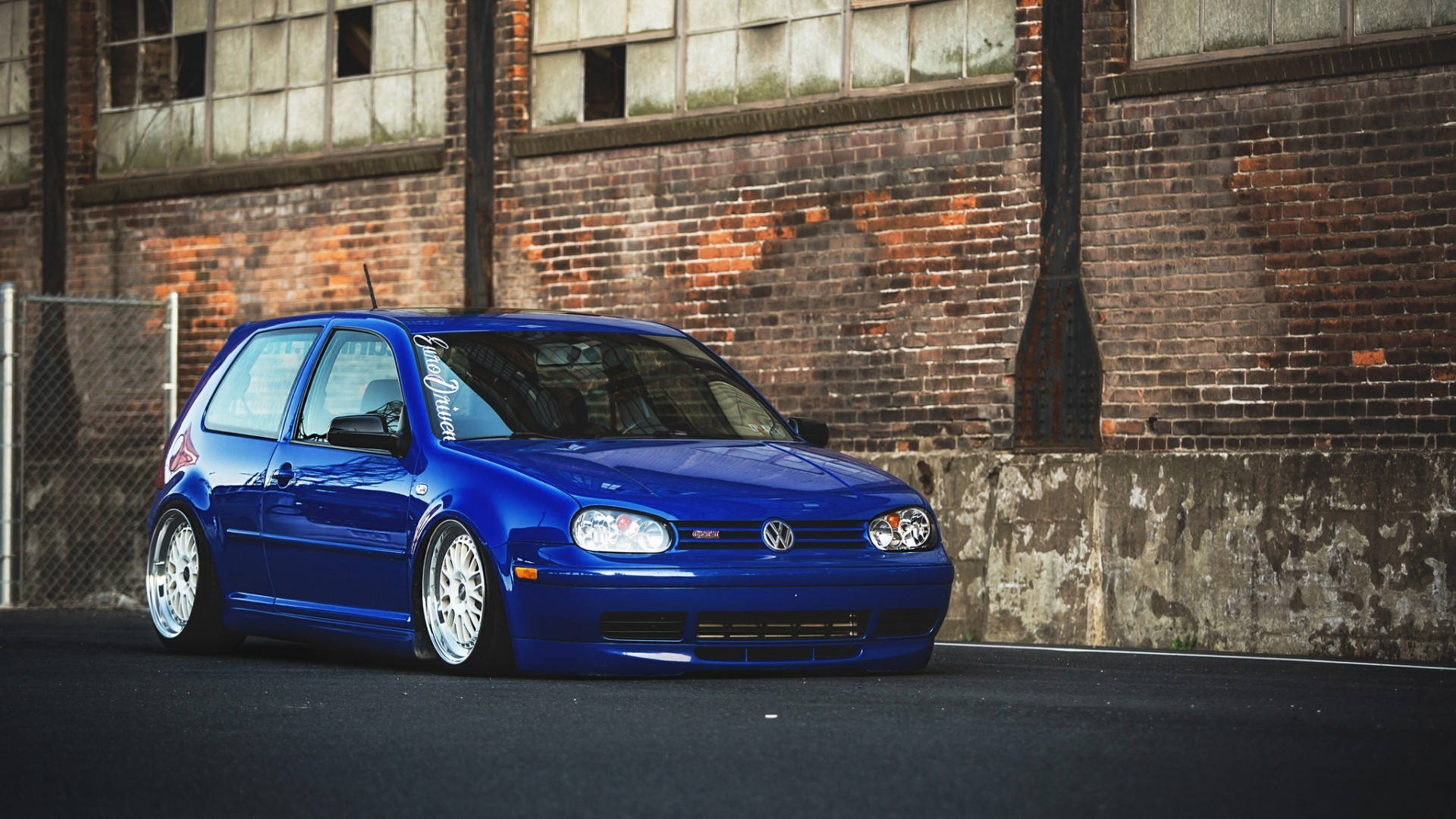 藍色Mk4 Volkswagen Golf GTI 壁紙 — 一輛鮮藍色改裝福士Golf低趴於白色輪圈上，背景是風化的磚牆倉庫。