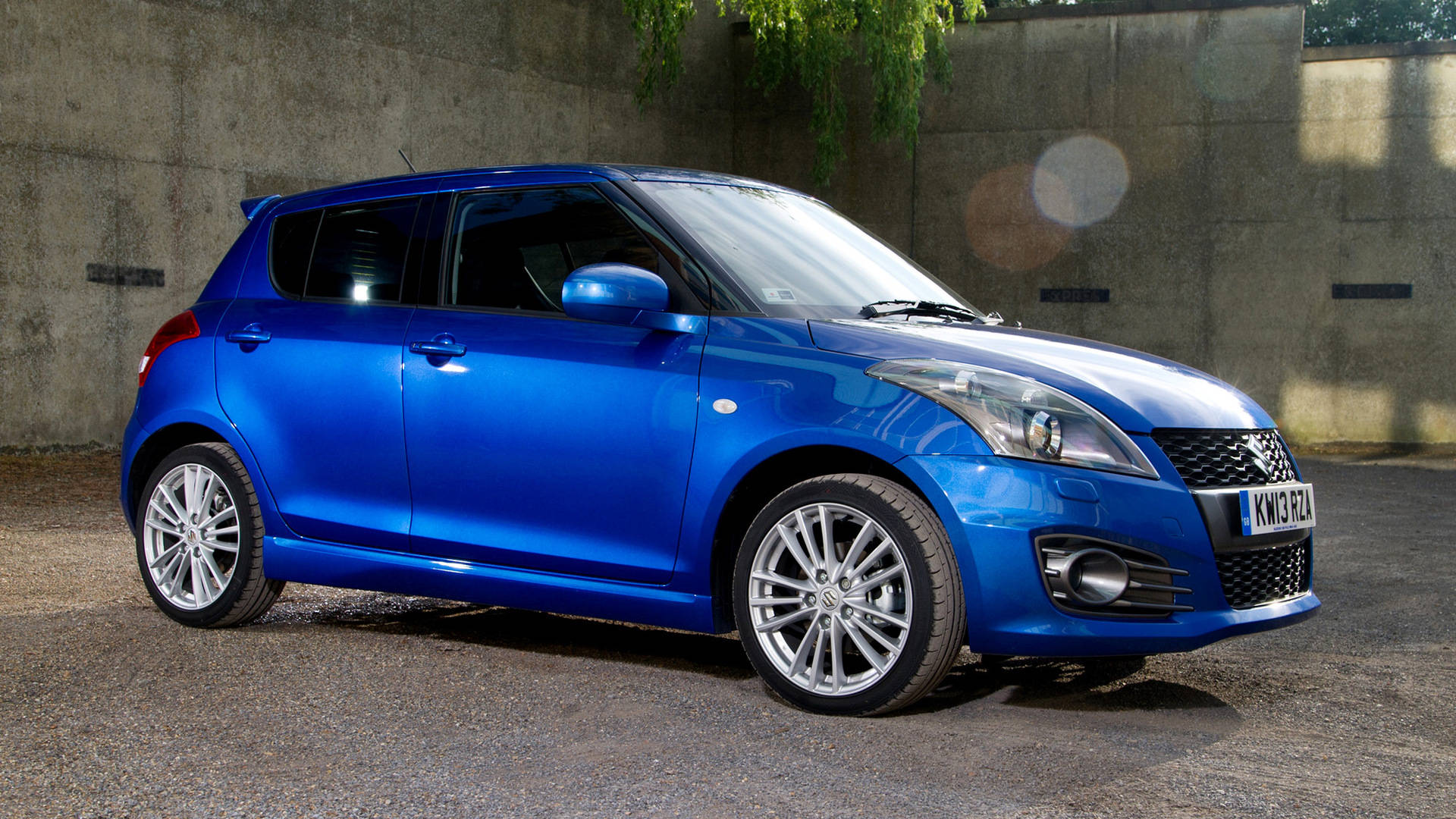 藍色Suzuki Swift 壁紙 — 一架充滿活力的藍色鈴木 Swift 停泊在碎石上，背景是帶紋理的混凝土牆，陽光斑駁，鏡頭眩光。