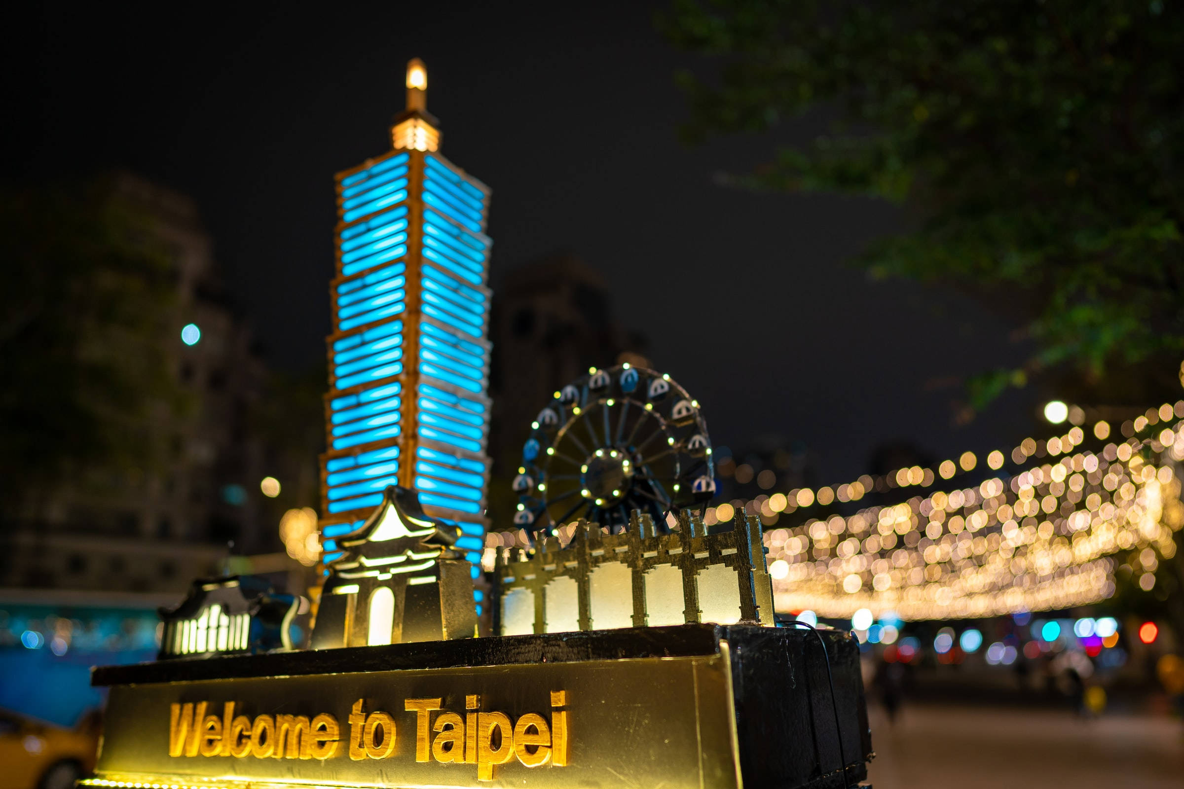 藍色台北101紀念品 壁紙 — 夜晚發光的「Welcome to Taipei」標誌，背景有藍色燈光的摩天大樓和摩天輪。