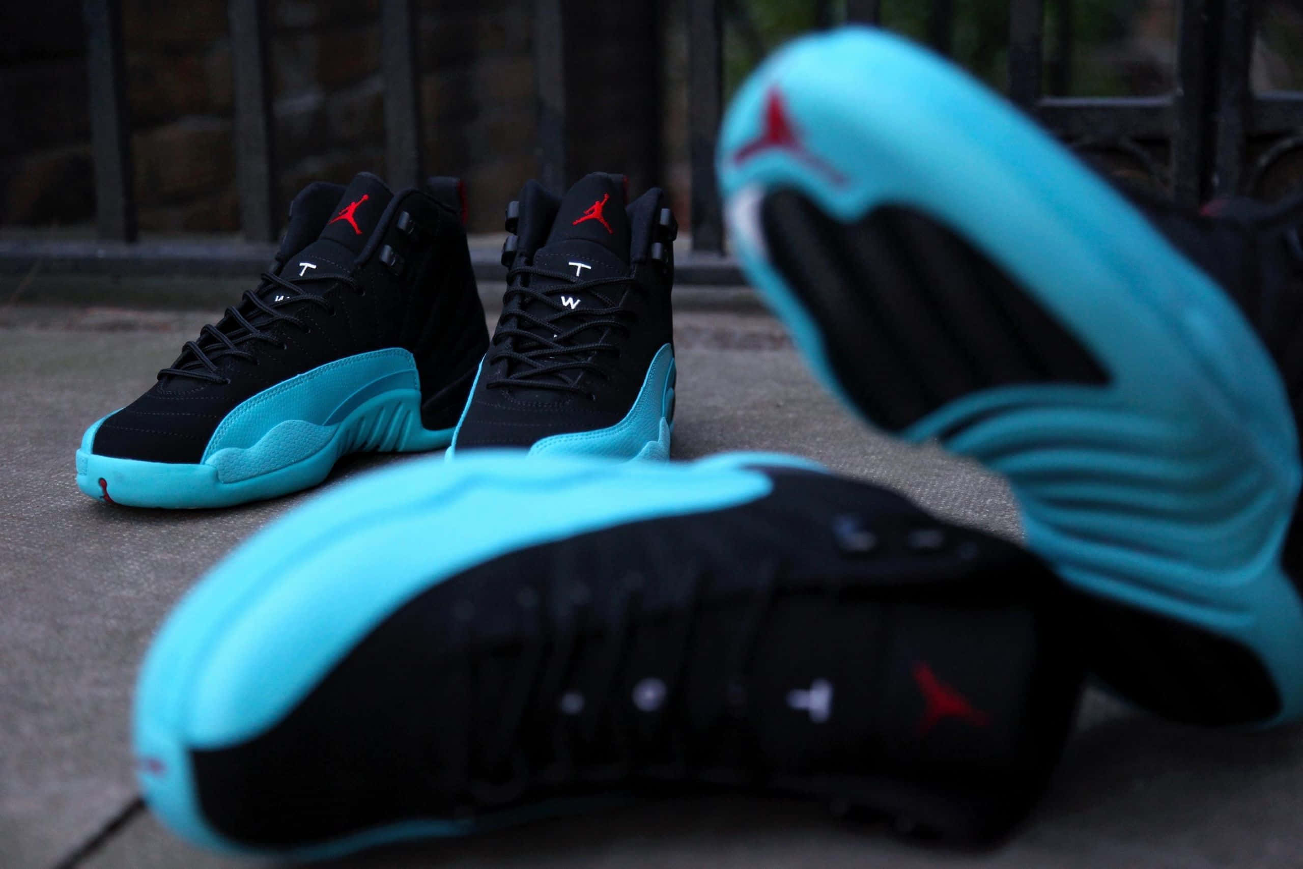 藍色喬丹 AJ Retro 12 壁紙 — 黑色和藍綠色的Jordan 12運動鞋帶有紅色標誌，排列在石頭庭院中，配有鍛鐵圍欄，在柔和日光下。