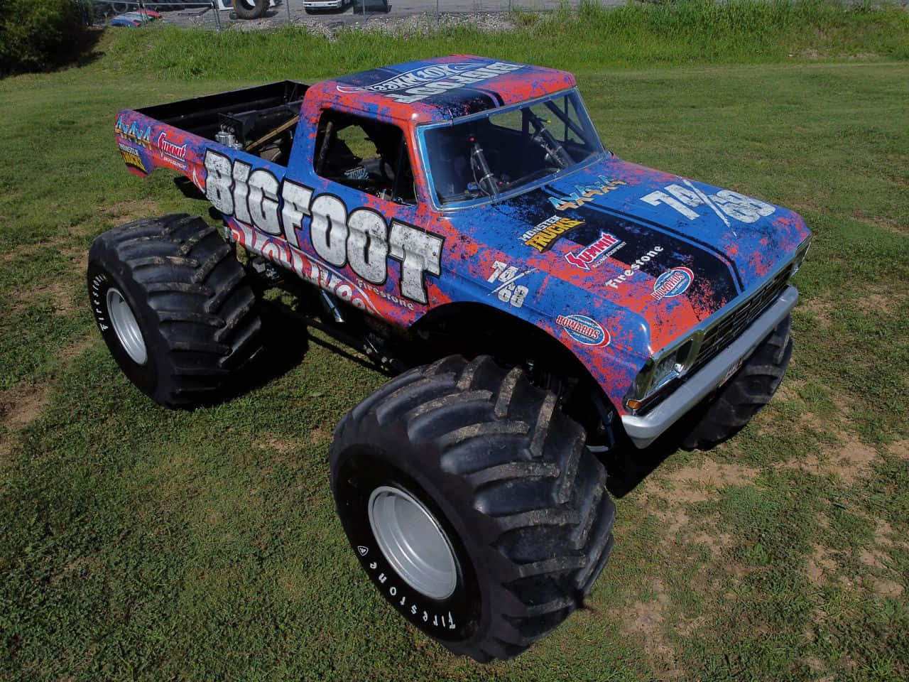 藍色大腳怪怪物卡車 壁紙 — 一輛名為「Bigfoot」的鮮豔藍紅怪獸卡車，配備巨大輪胎，停在陽光明媚的草地上。