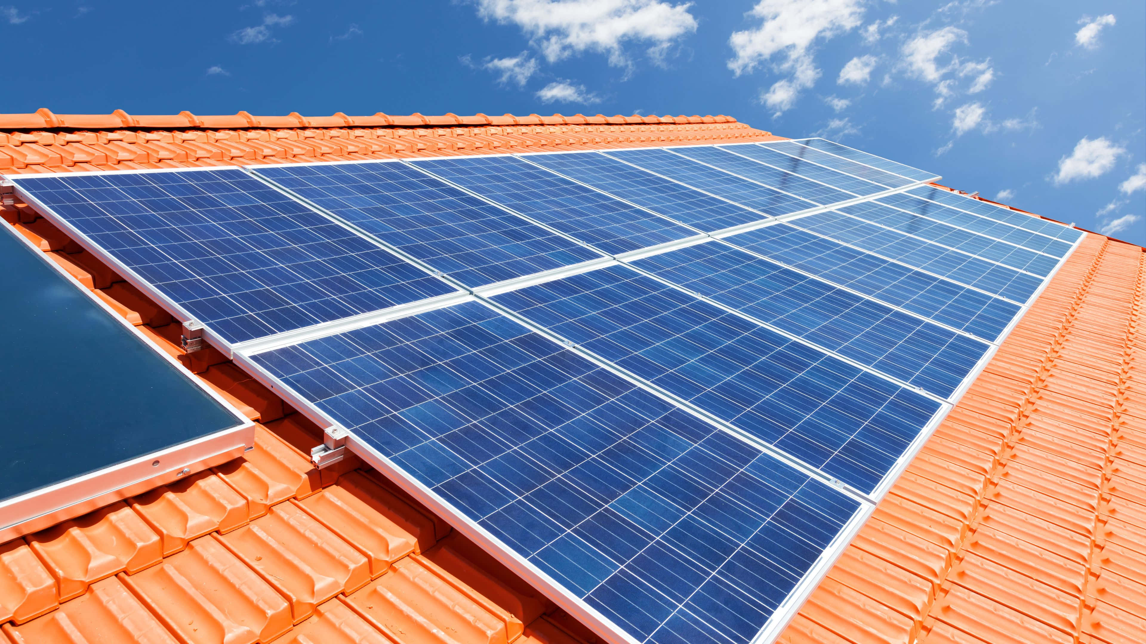 Solar Panel 桌布 壁紙 — 藍色太陽能板在赤陶屋頂上閃閃發光，天空湛藍，白雲點點。