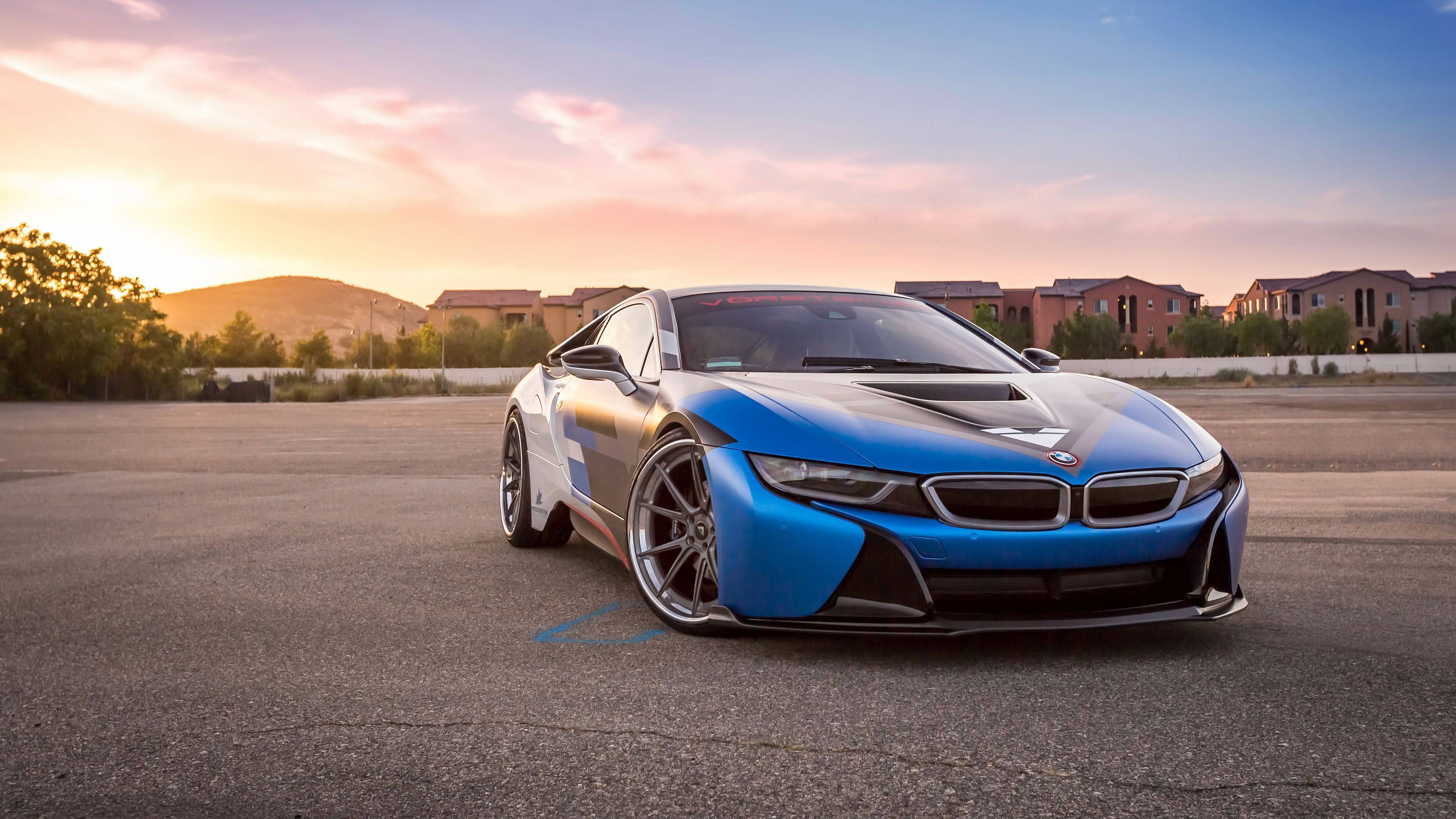 藍色寶馬車後的日出 壁紙 — 一架時尚的藍色BMW i8跑車停在夕陽下的瀝青路上，背景有溫暖的天空和遠處的房屋。