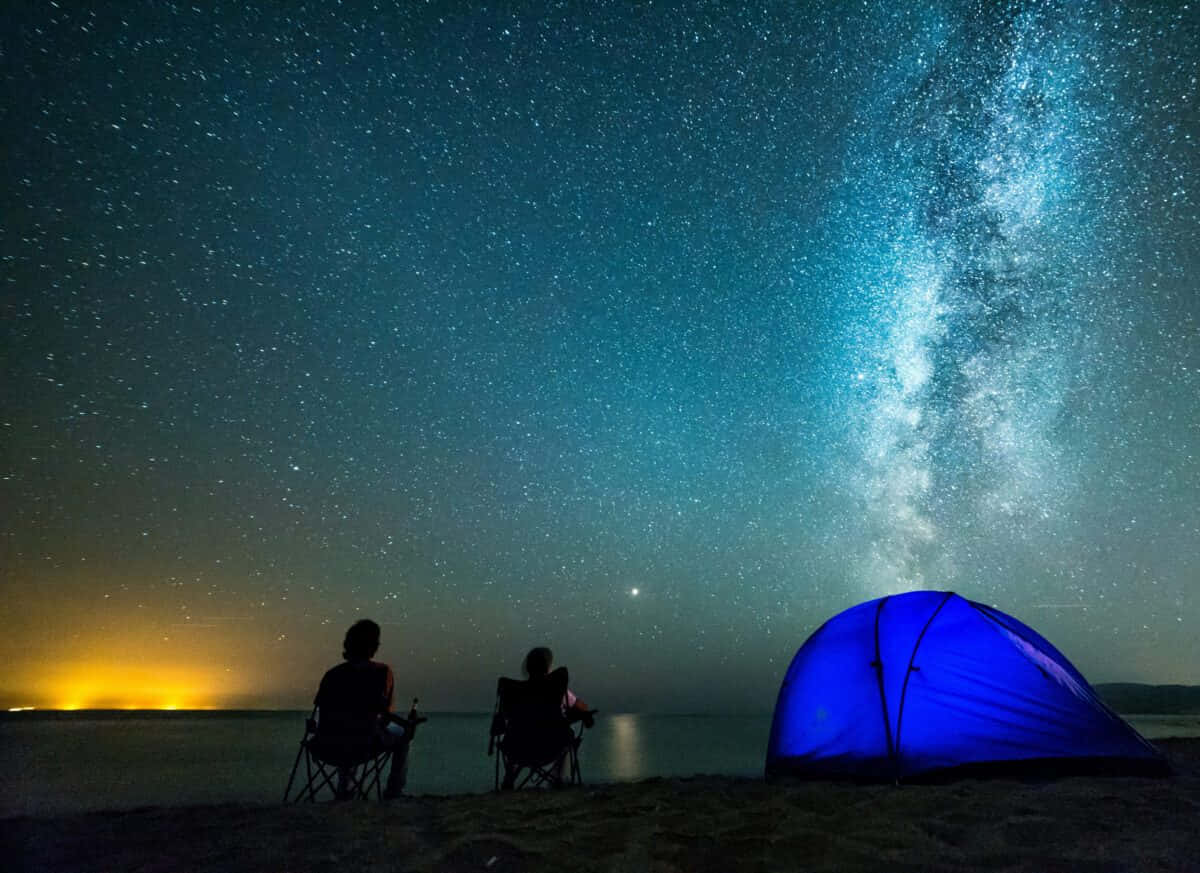 兩個人坐在椅子上，旁邊是一個藍色帳篷，在星空下，銀河清晰可見。