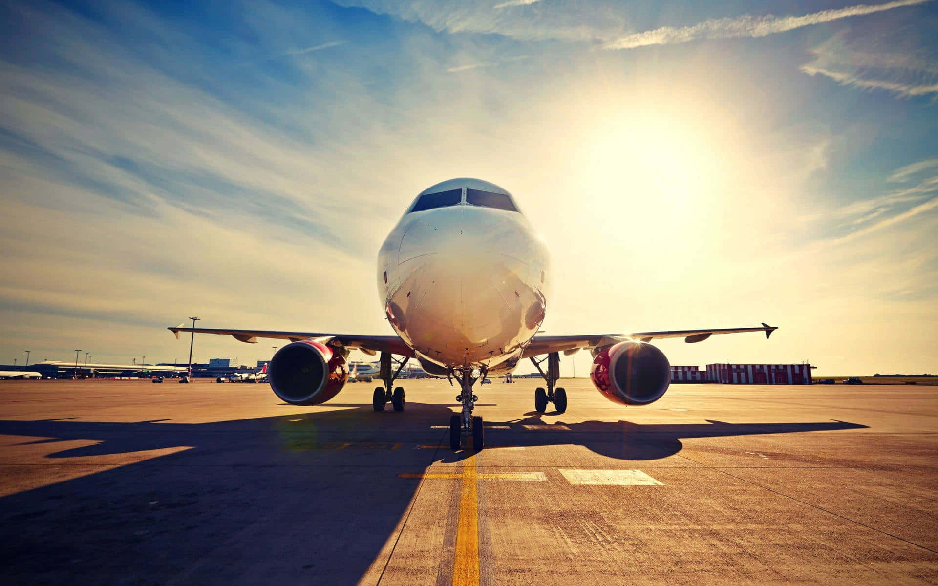 藍色日落天空飛機背景低角度拍攝 壁紙 — 一架商用飛機面向鏡頭，停在陽光普照的停機坪上，機頭後方閃耀著金色光芒。