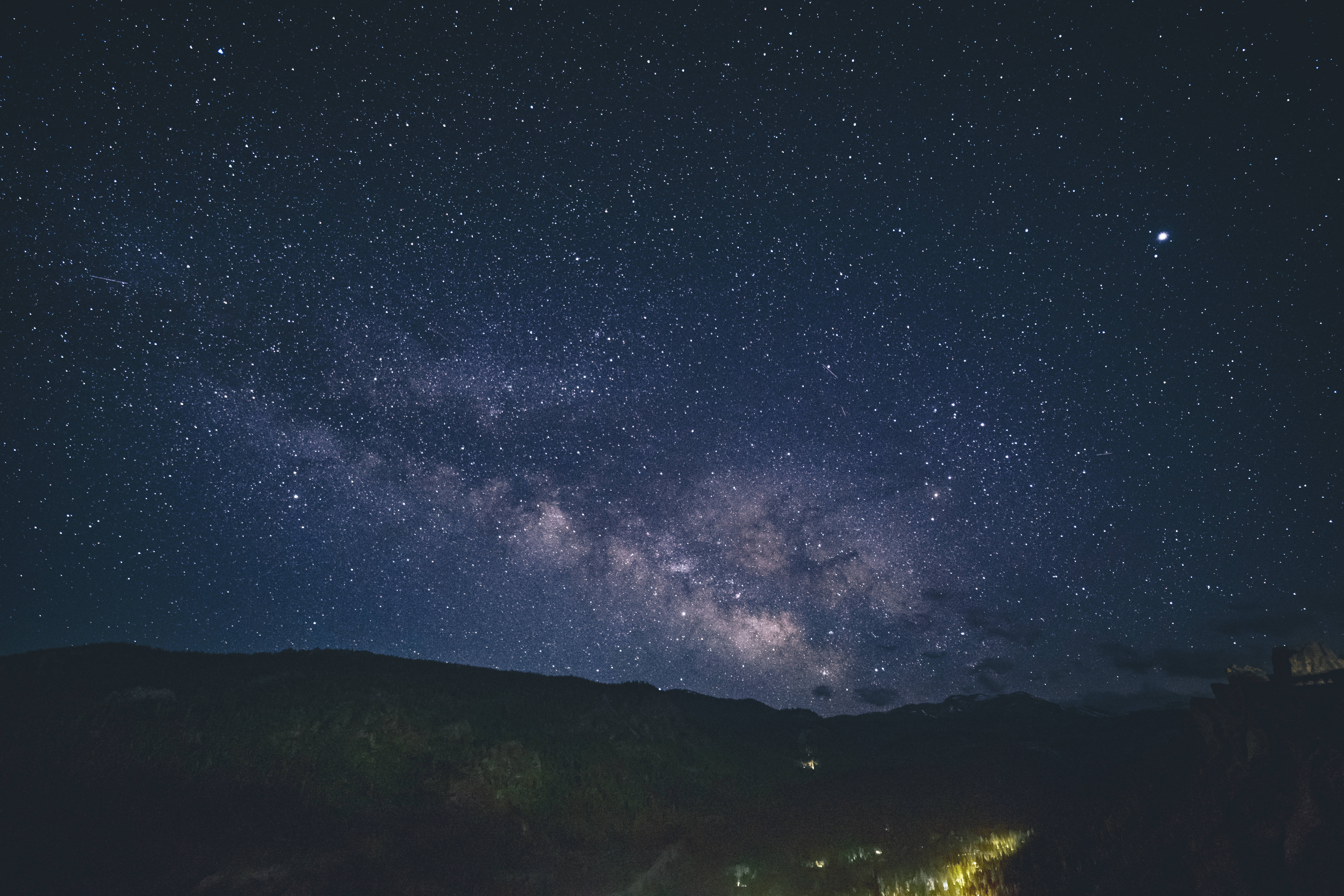 藍色星系星夜風景 壁紙 — 令人嘆為觀止的夜空，充滿無數星星，發光的銀河在黑暗的山脈上方拱起，下方有遙遠的村莊燈光。