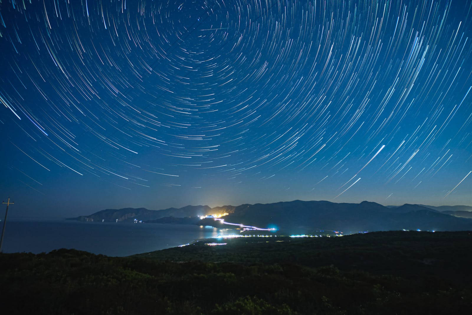 藍色星系繁星夜空 壁紙 — 星軌在深藍色夜空中盤旋，下方是沿海小鎮，海岸線上有閃爍的燈光。