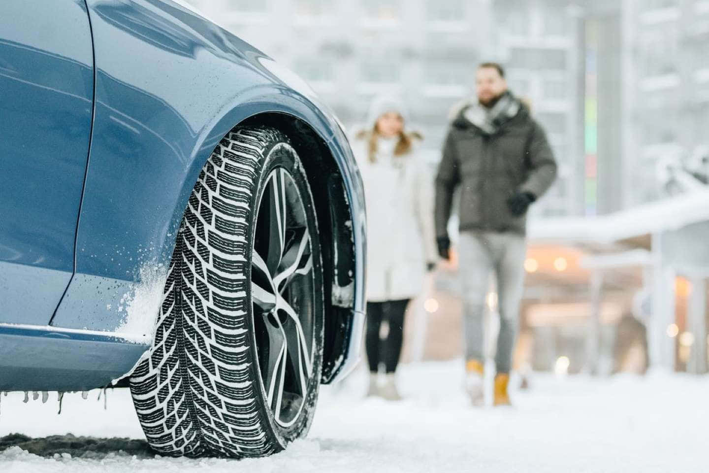 Winter Tires 桌布 壁紙 — 藍色汽車輪胎被雪覆蓋的特寫，背景有一對情侶在雪中行走。
