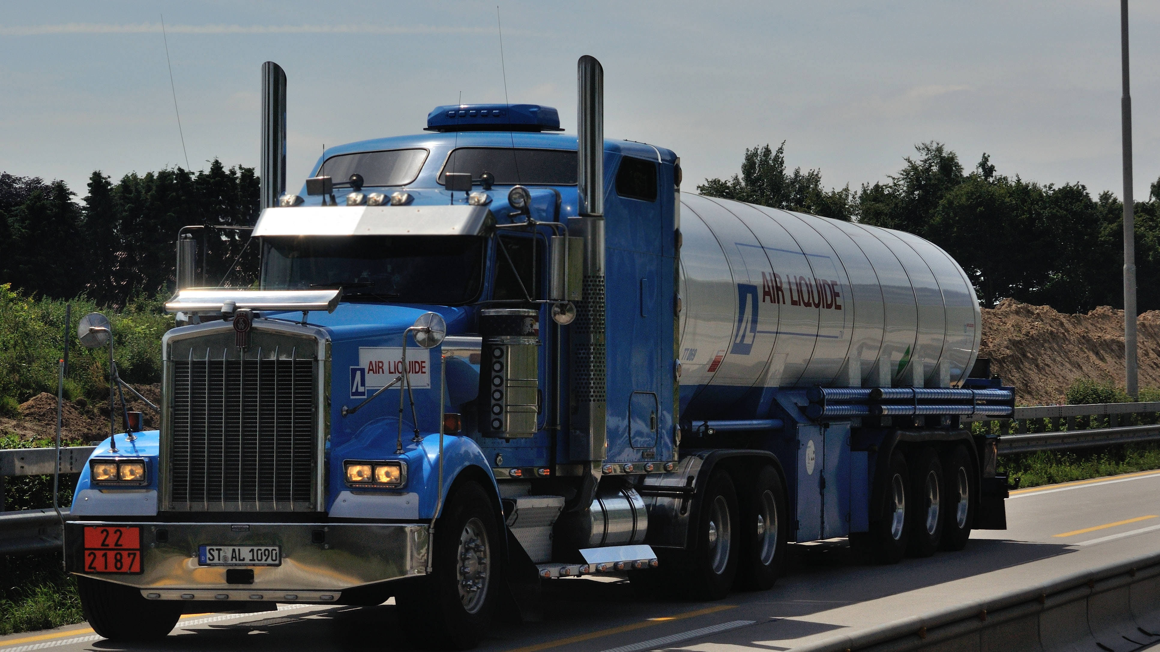 藍色油罐車肯沃斯W900 壁紙 — 一輛藍色 Air Liquide 油罐車在高速公路上行駛，背景有綠樹和晴朗的天空。