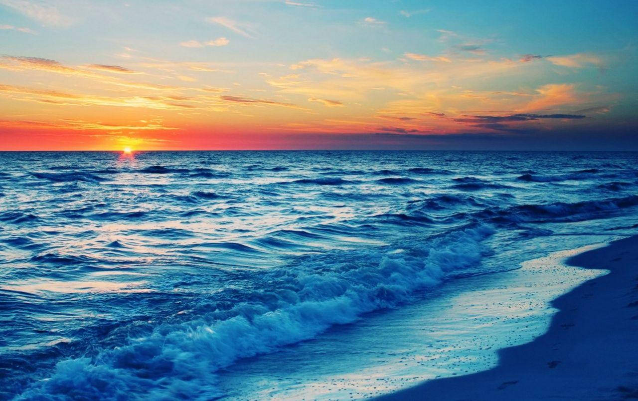 藍色海浪拍打海灘日落 壁紙 — 夕陽將天空染成火熱的橙色和粉紅色，海浪在廣闊的海洋下拍打著沙灘。