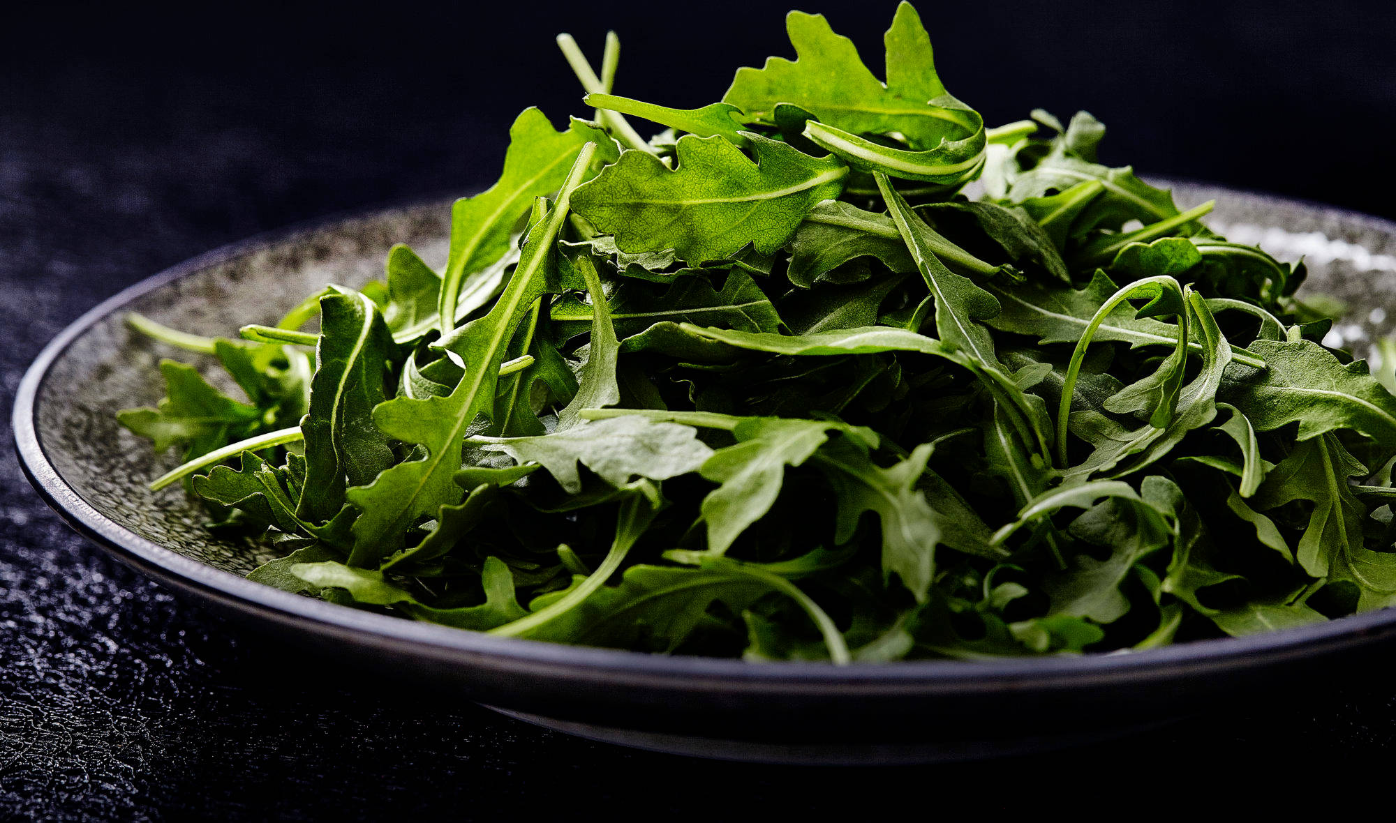 藍色湯盤上的芝麻菜葉堆 壁紙 — 特寫新鮮翠綠的芝麻菜葉，高高堆疊在一個深色紋理陶瓷碗中，背景為黑色。