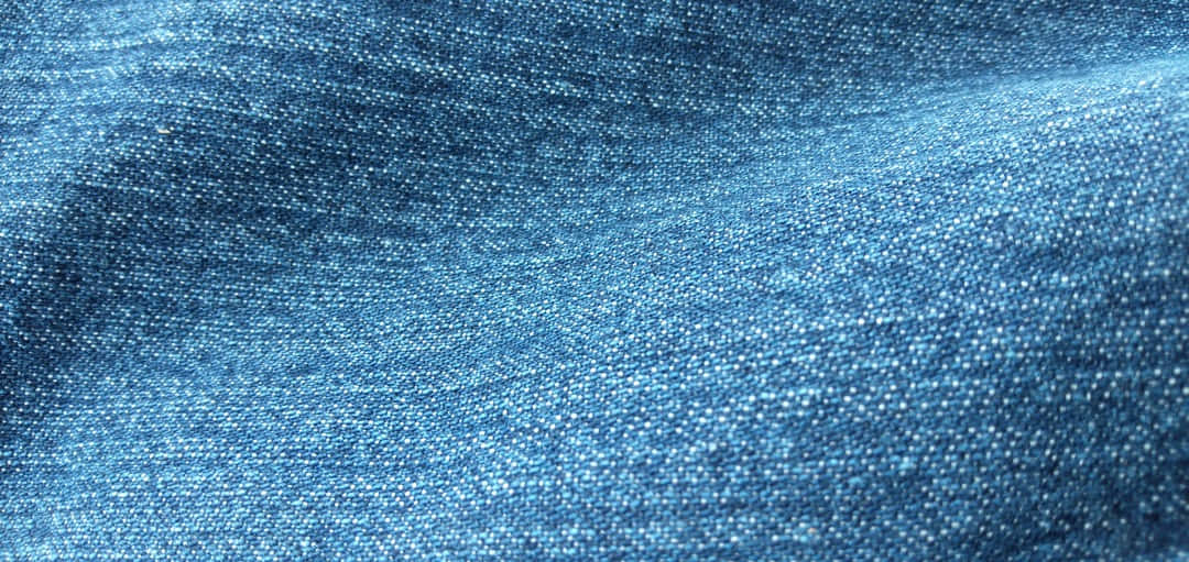 Denim Blue 桌布 壁紙 — 藍色紋理牛仔布料的特寫，在均勻光線下可見編織紋理和柔軟摺痕。