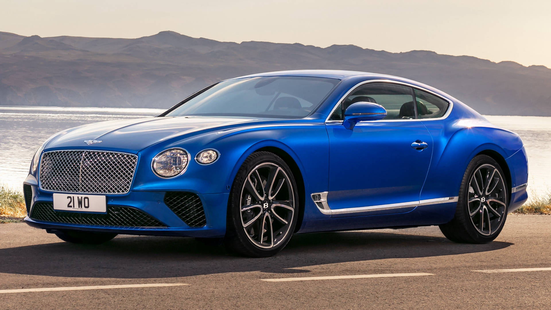 日落時分，藍色 Bentley Continental GT 停泊在水邊，背景有山脈。