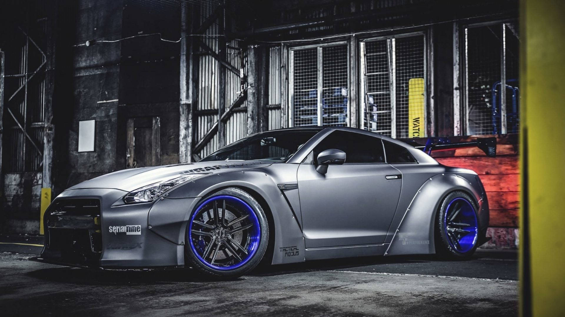 定制銀色日產GT-R，配備藍色輪圈，停泊在黑暗的工業倉庫中。