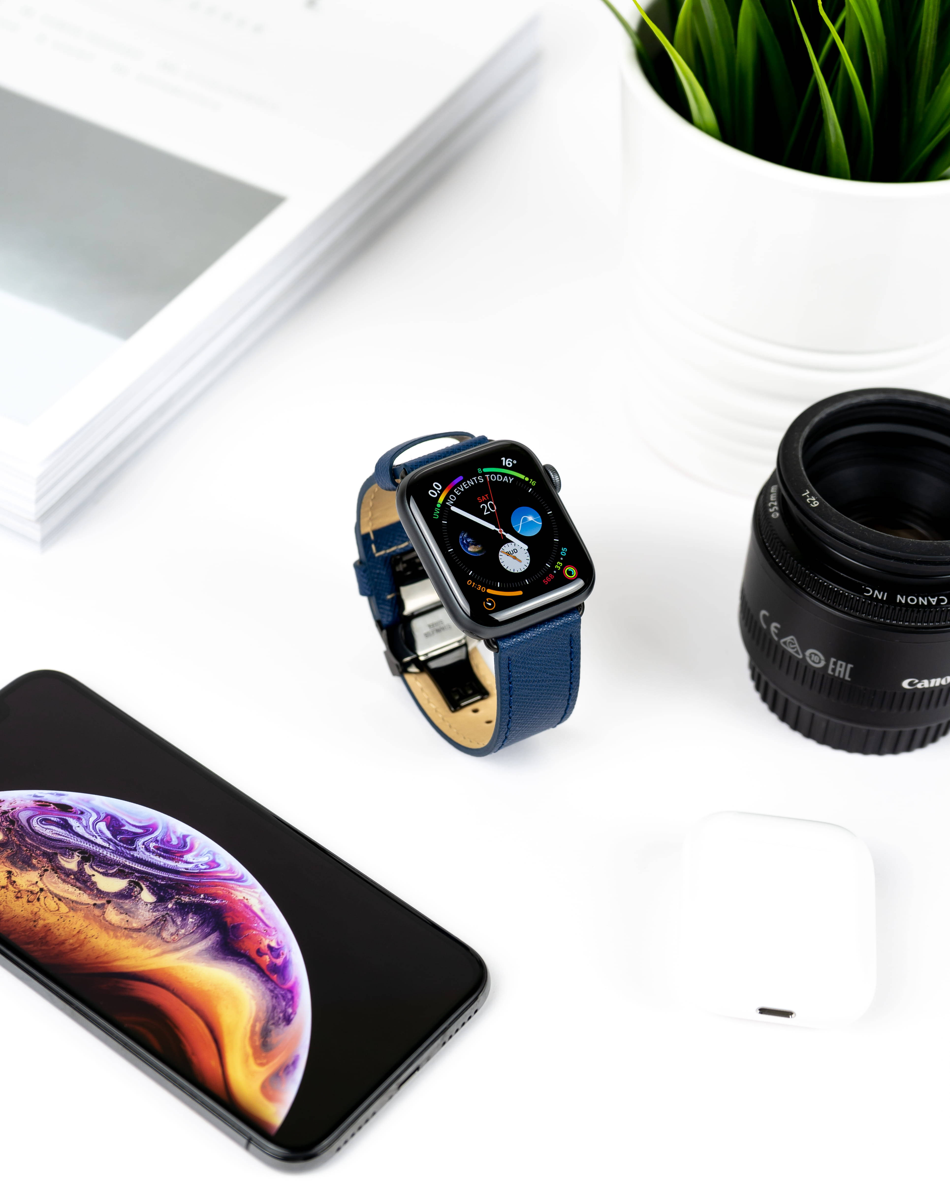 藍色錶帶 Apple Watch 4K iPhone 11 壁紙 — 一款時尚的智能手錶、手機相機鏡頭和AirPods放在簡約白色桌子上，旁邊有一盆植物和一本打開的書。