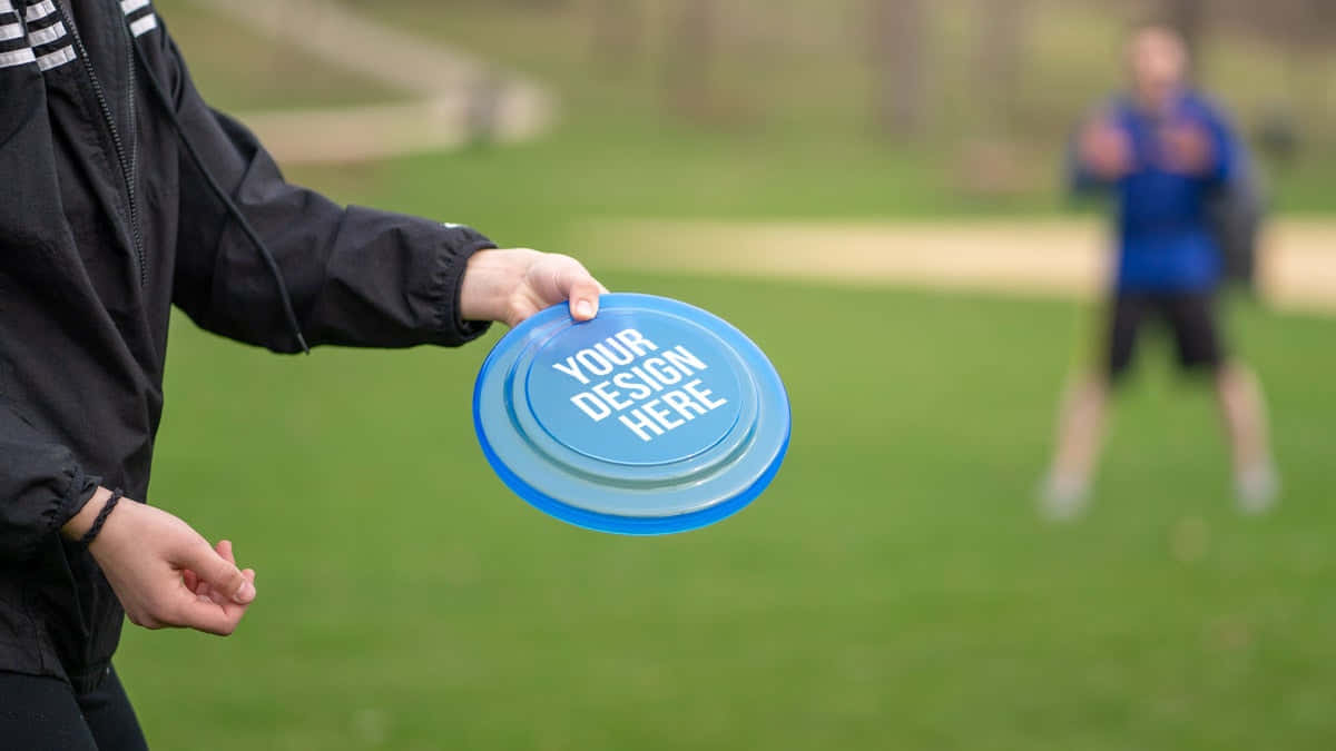 藍色飛盤模板最佳 Ultimate Frisbee 背景 壁紙 — 一個穿著黑色外套的人拿著一個有文字的藍色飛盤，準備在草地上拋出，背景有另一個模糊的玩家。