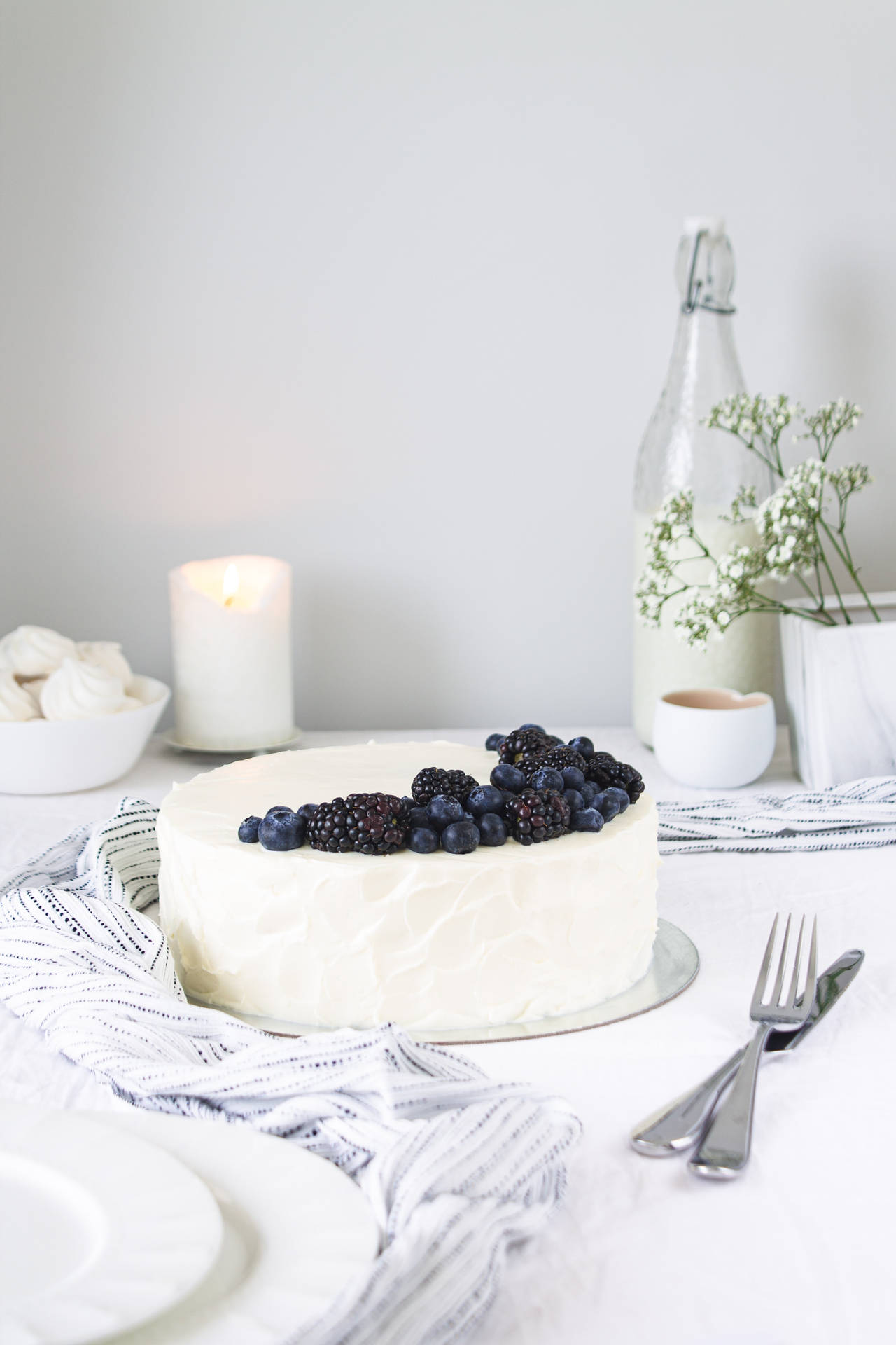 白色糖霜蛋糕配莓果，放在條紋布上，旁邊有蠟燭和玻璃瓶。