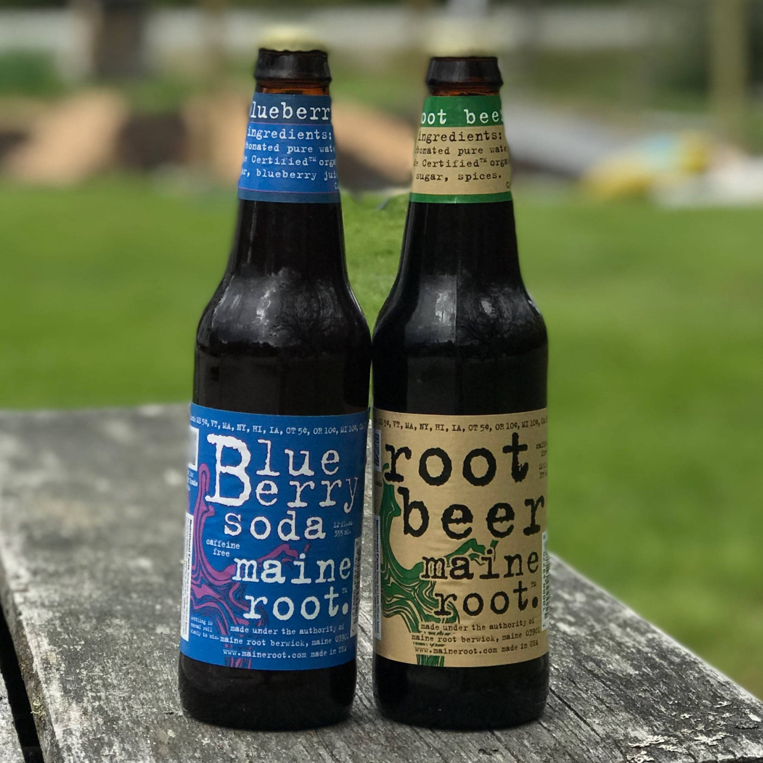 藍莓蘇打與根汁飲料 Maine 系列 壁紙 — 兩瓶深色玻璃瓶裝的Maine Root汽水和根汁啤酒並排放置在風化的木製野餐桌上，背景是模糊的綠色草坪。