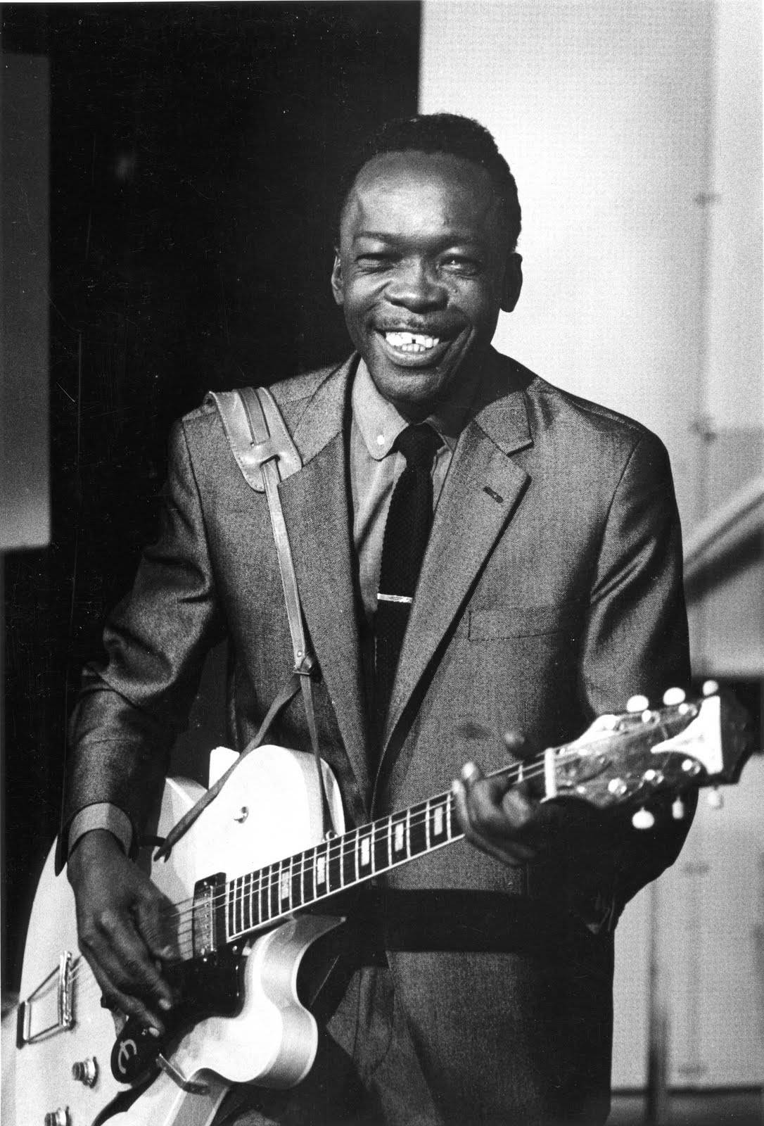 藍調傳奇 John Lee Hooker - 單色肖像照 壁紙 — 一位快樂的黑人男士穿著西裝，在工作室中拿著白色電結他，笑容燦爛。